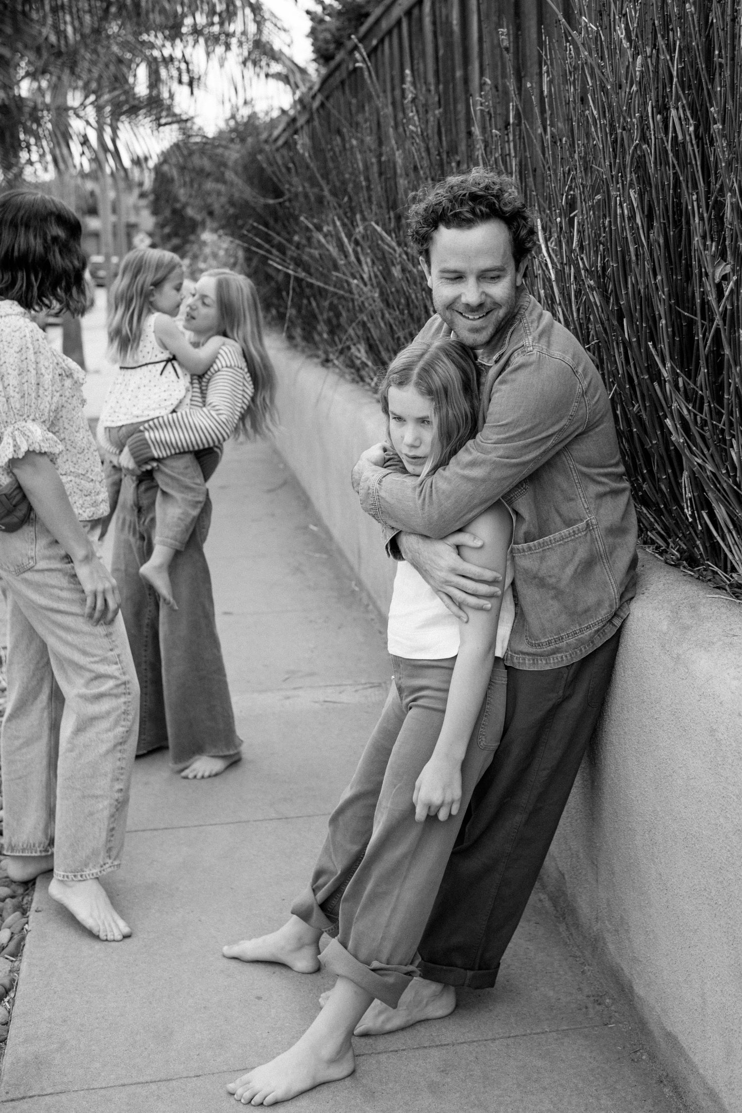 A black and white photo of a curly-haired father hugging his daughter against a wall while family stands nearby.
