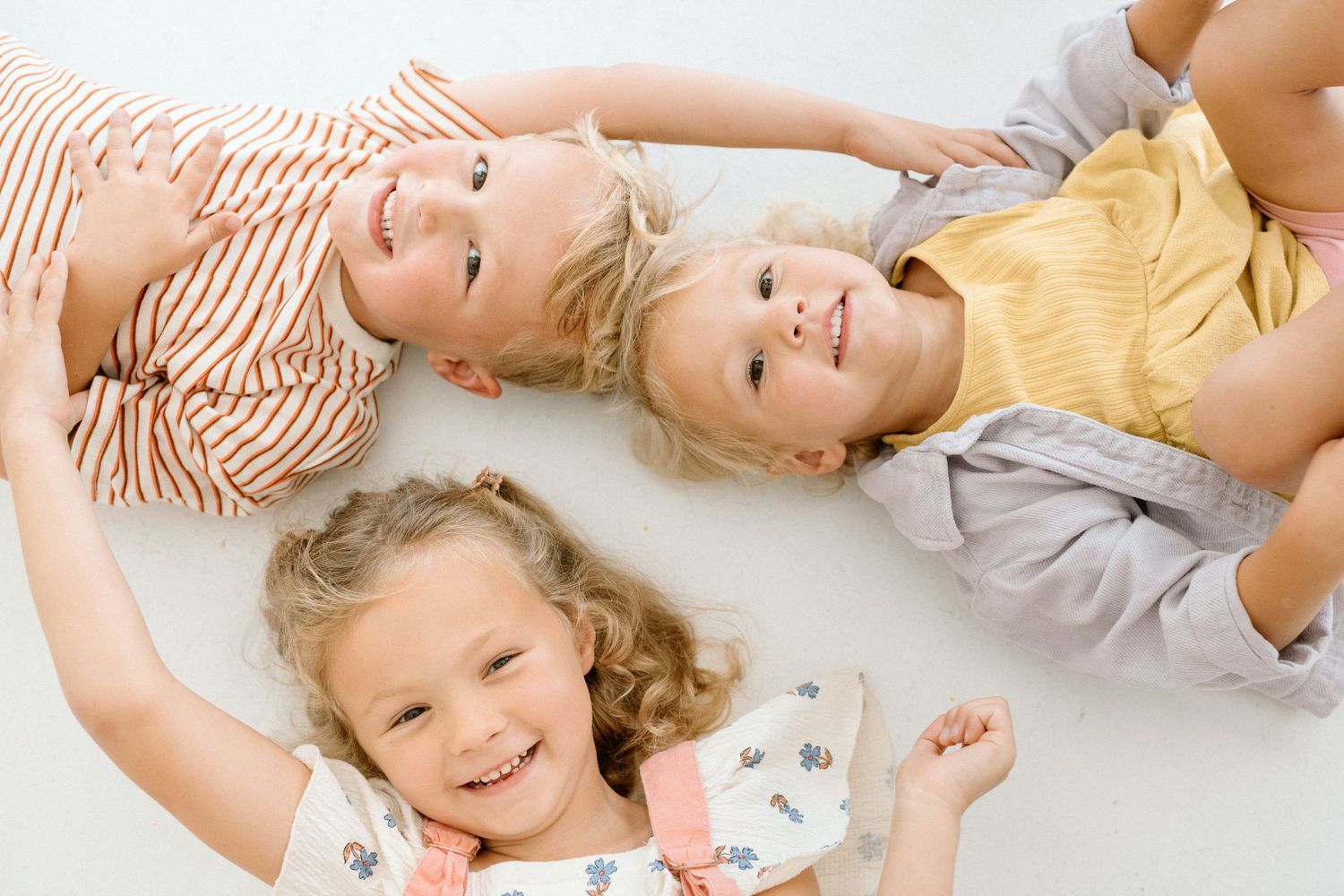 Three young blonde children lying on floor smiling up at camera in cheerful portrait.