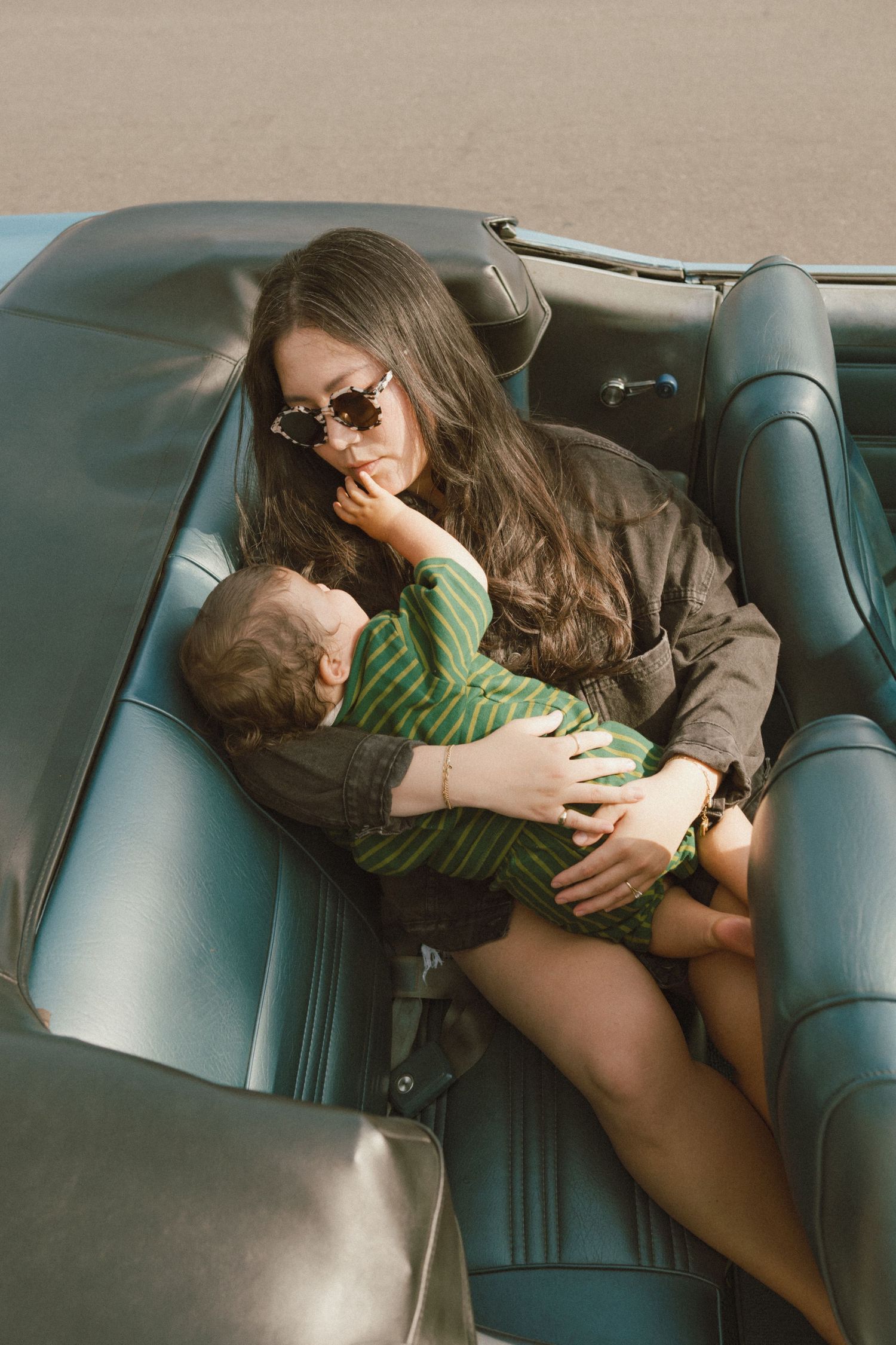 Young woman with sunglasses holding a baby in a vintage convertible car with teal leather interior.