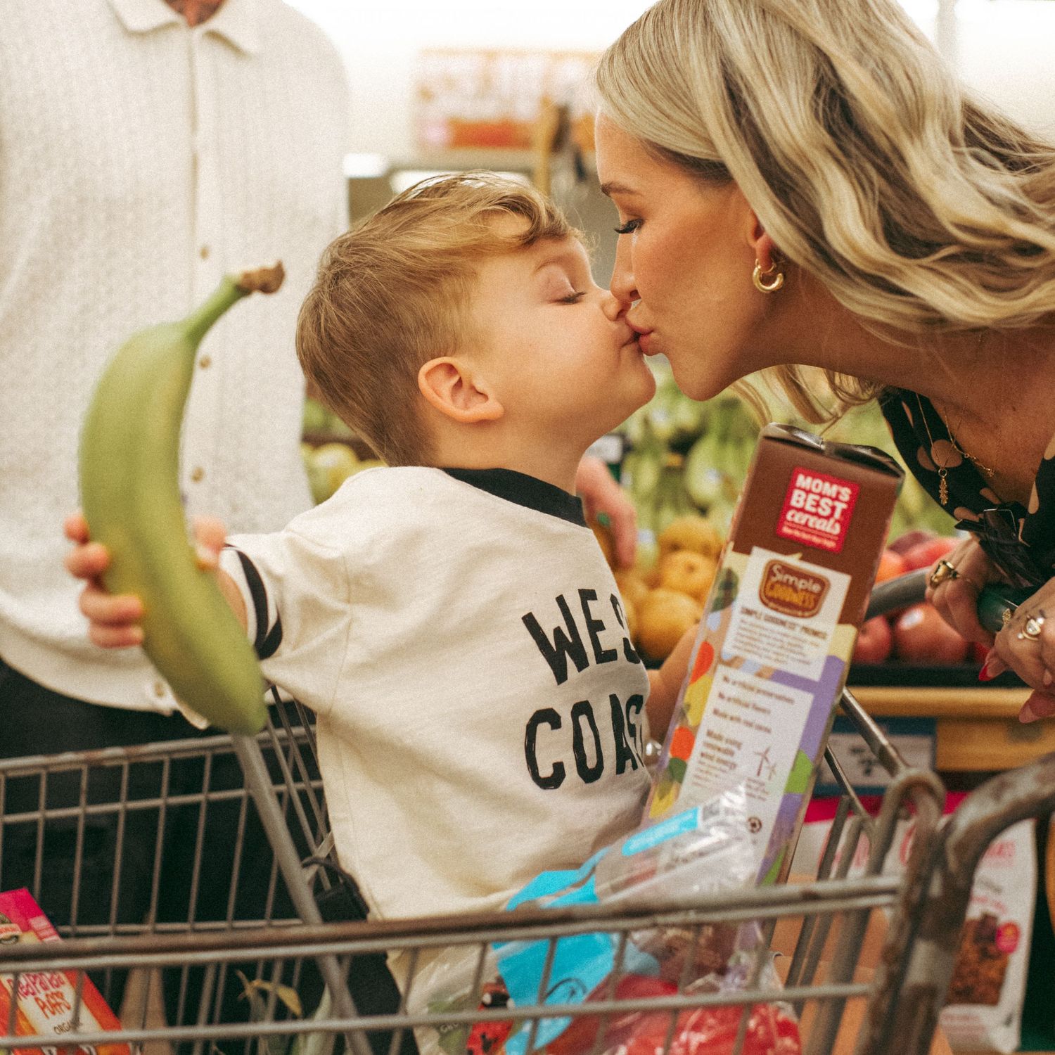 A mother kisses her toddler son sitting in a grocery cart holding a banana inside a supermarket.