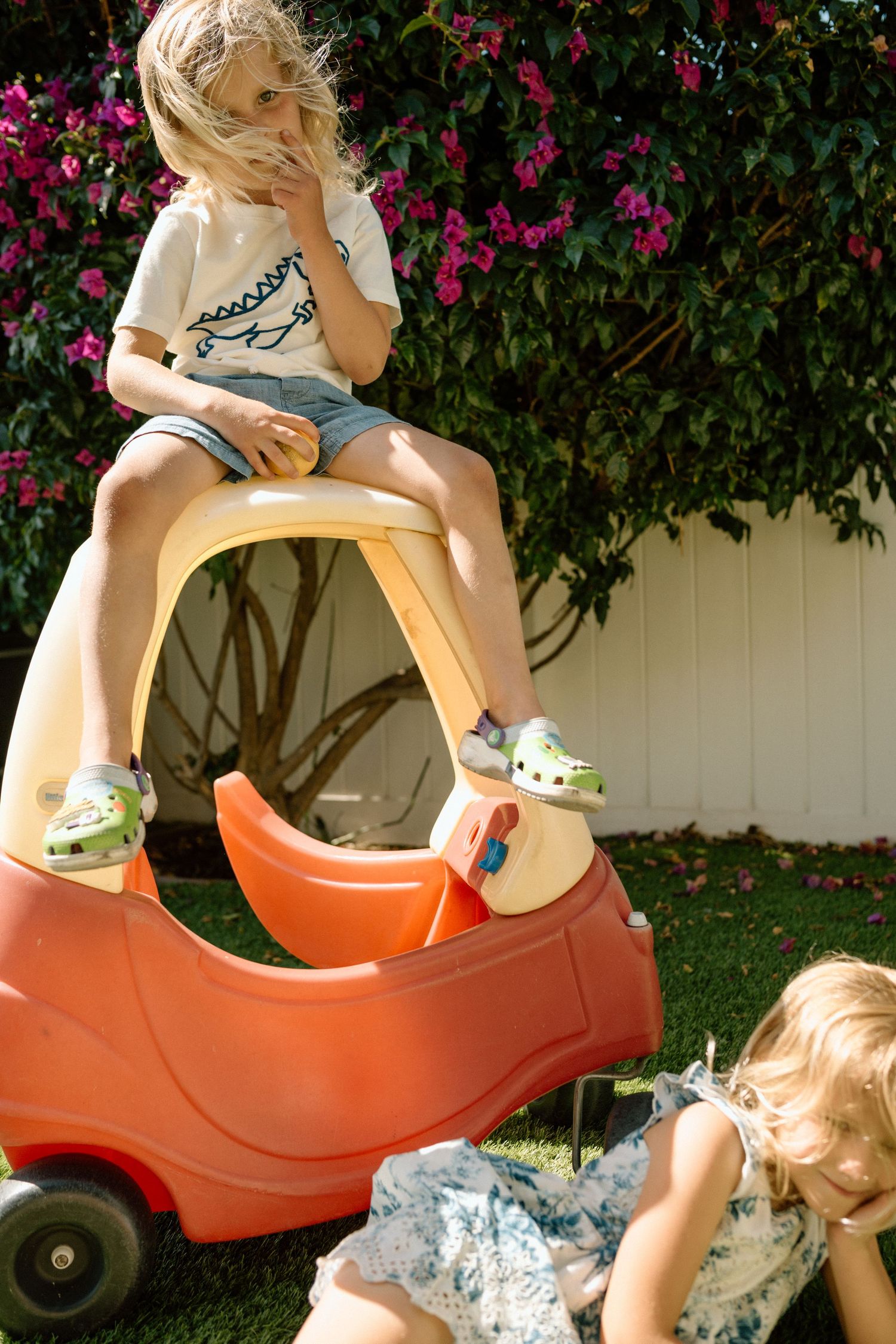 Two young children playing outdoors beside a colorful Little Tikes toy car with pink flowers in the background.