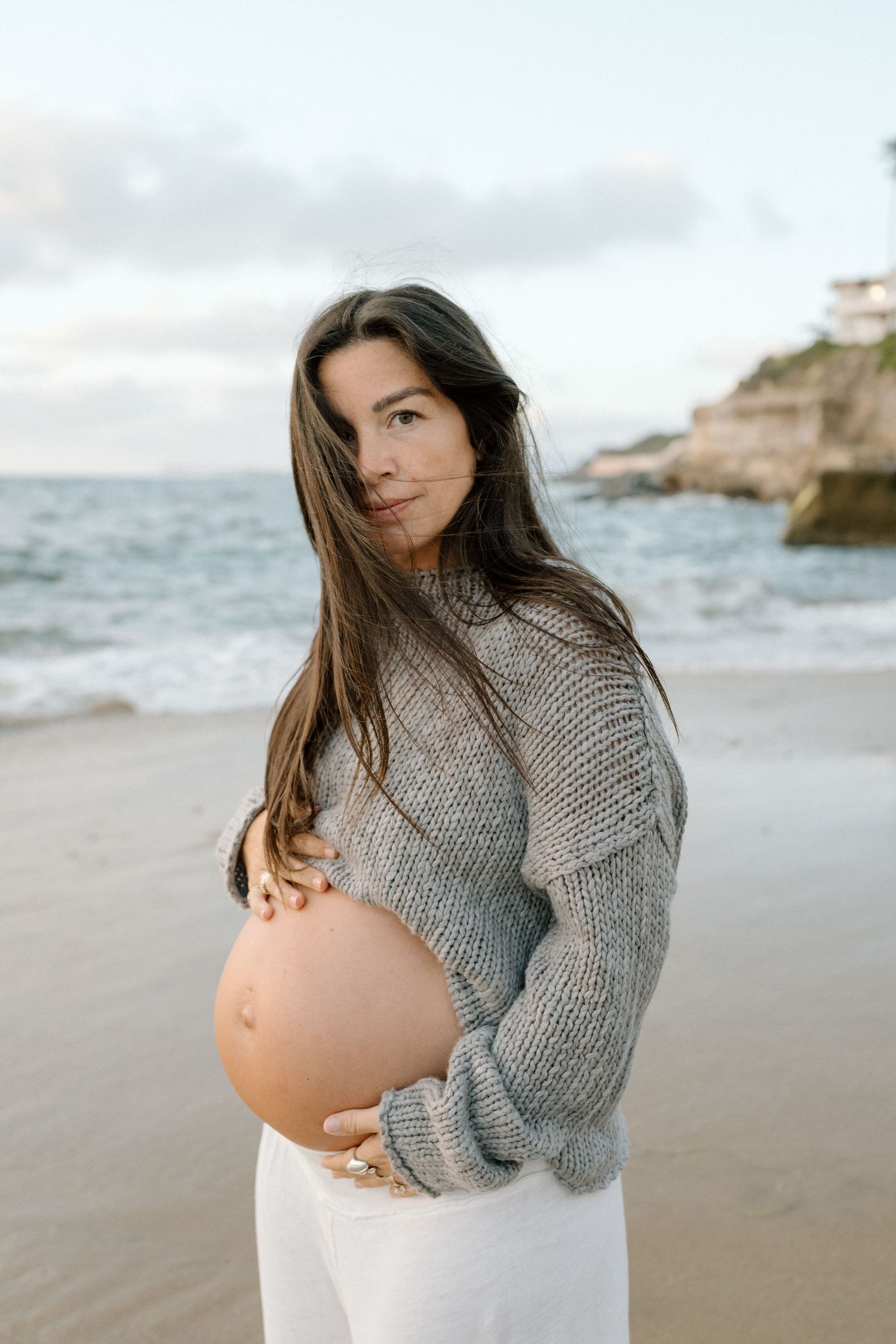 Pregnant woman in grey sweater cradling baby bump on a scenic beach at sunset.