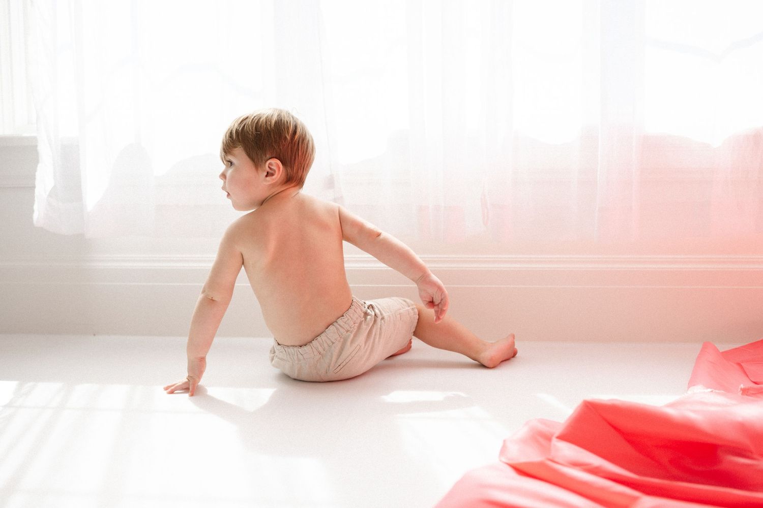 Toddler boy sitting on white floor in diaper, back to camera, in bright airy room with pink fabric nearby.