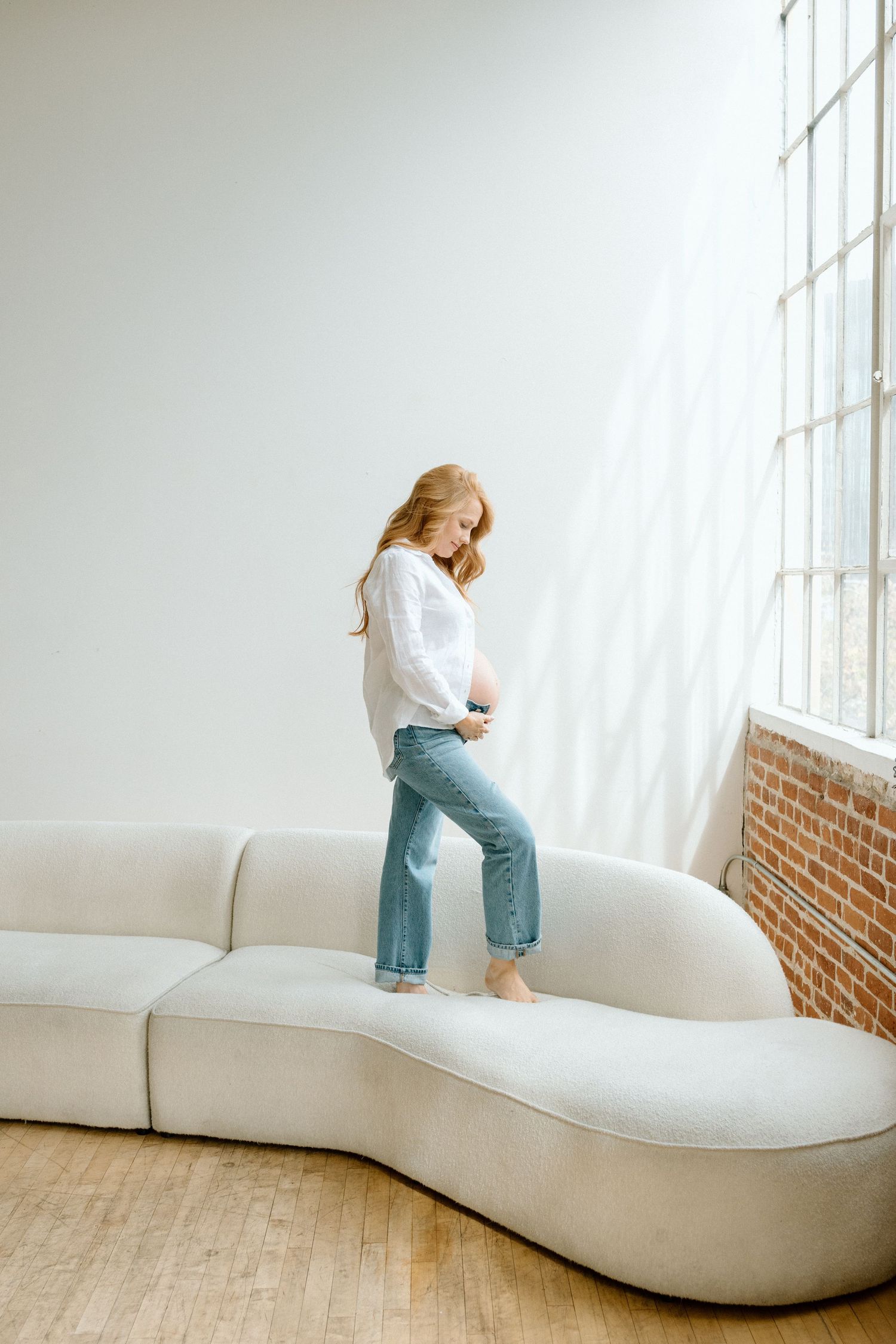 Pregnant woman in white shirt and jeans standing on a curved sofa near a window.