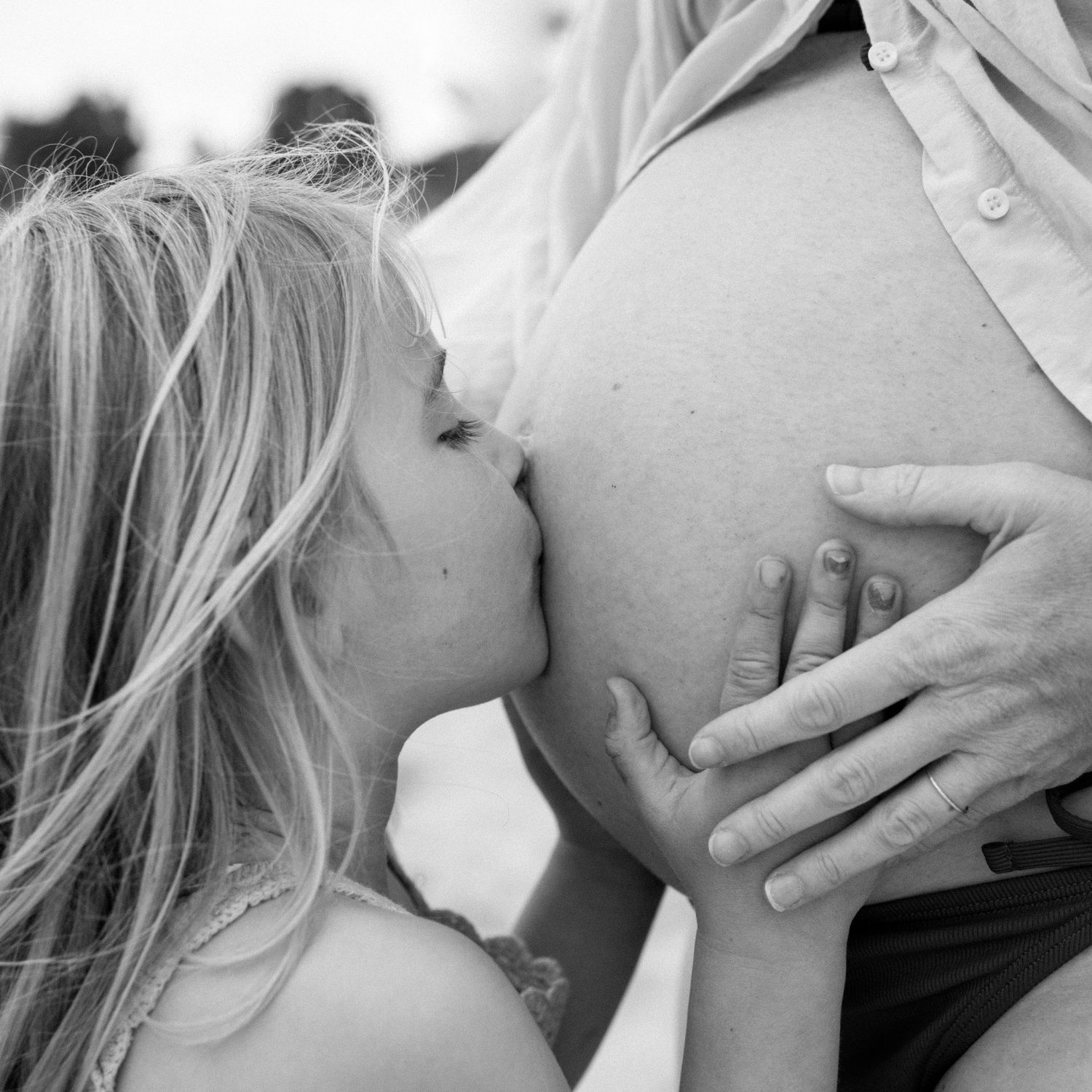 Black and white close-up of young blonde girl kissing pregnant mothers bare baby bump tenderly.