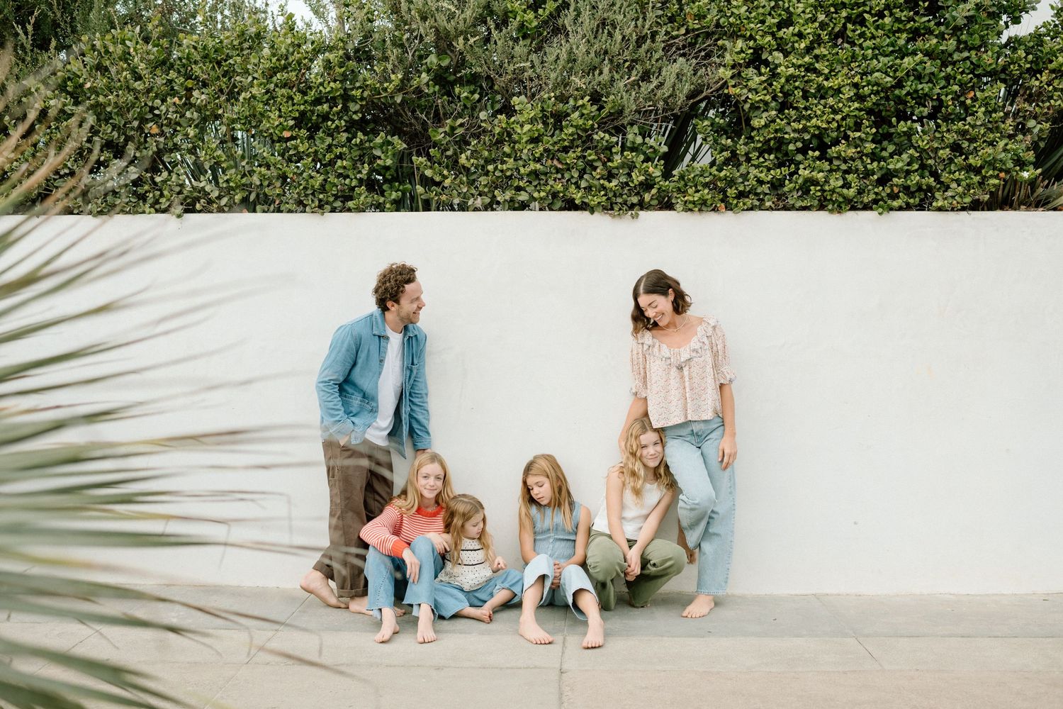 A family of six with parents and four young daughters relaxing against a white outdoor wall surrounded by greenery.