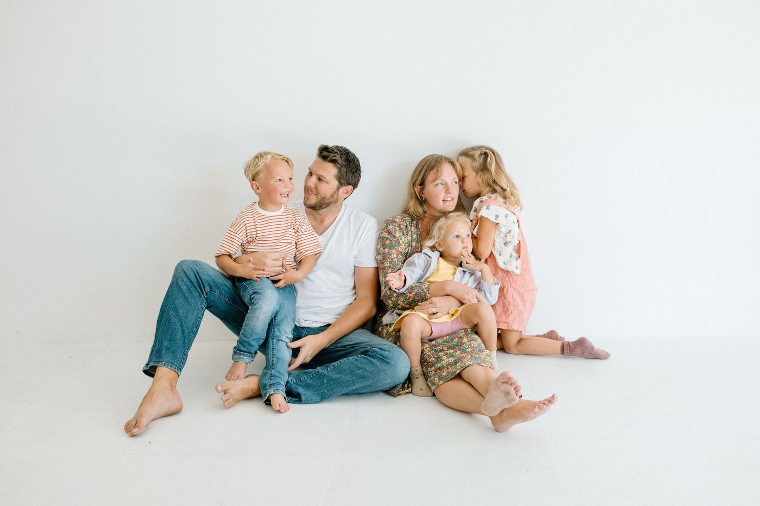 Happy family of five sitting together on white studio floor in casual warm-toned outfits.