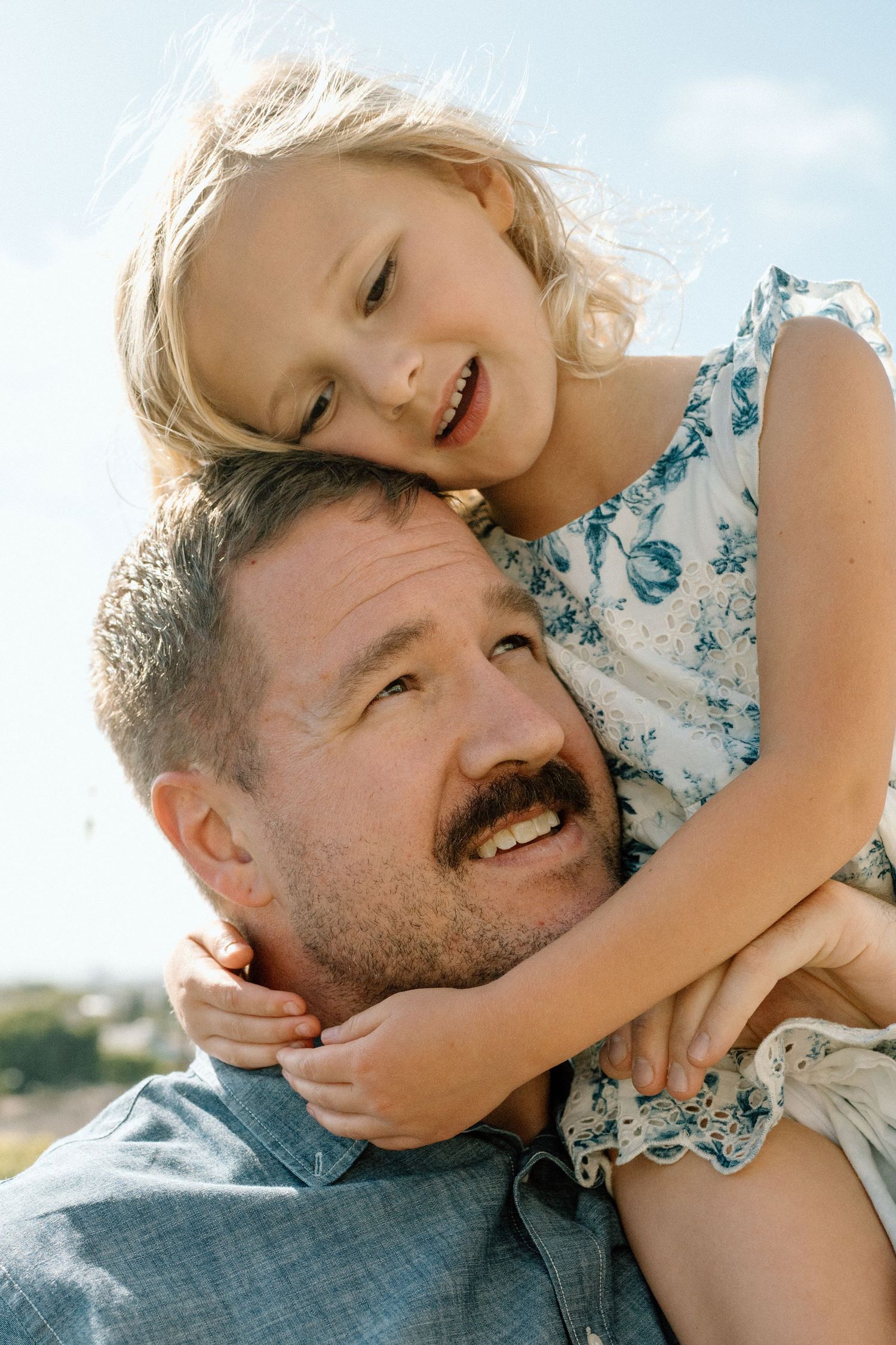A smiling young girl hugs a bearded man from behind outdoors against a bright blue sky.