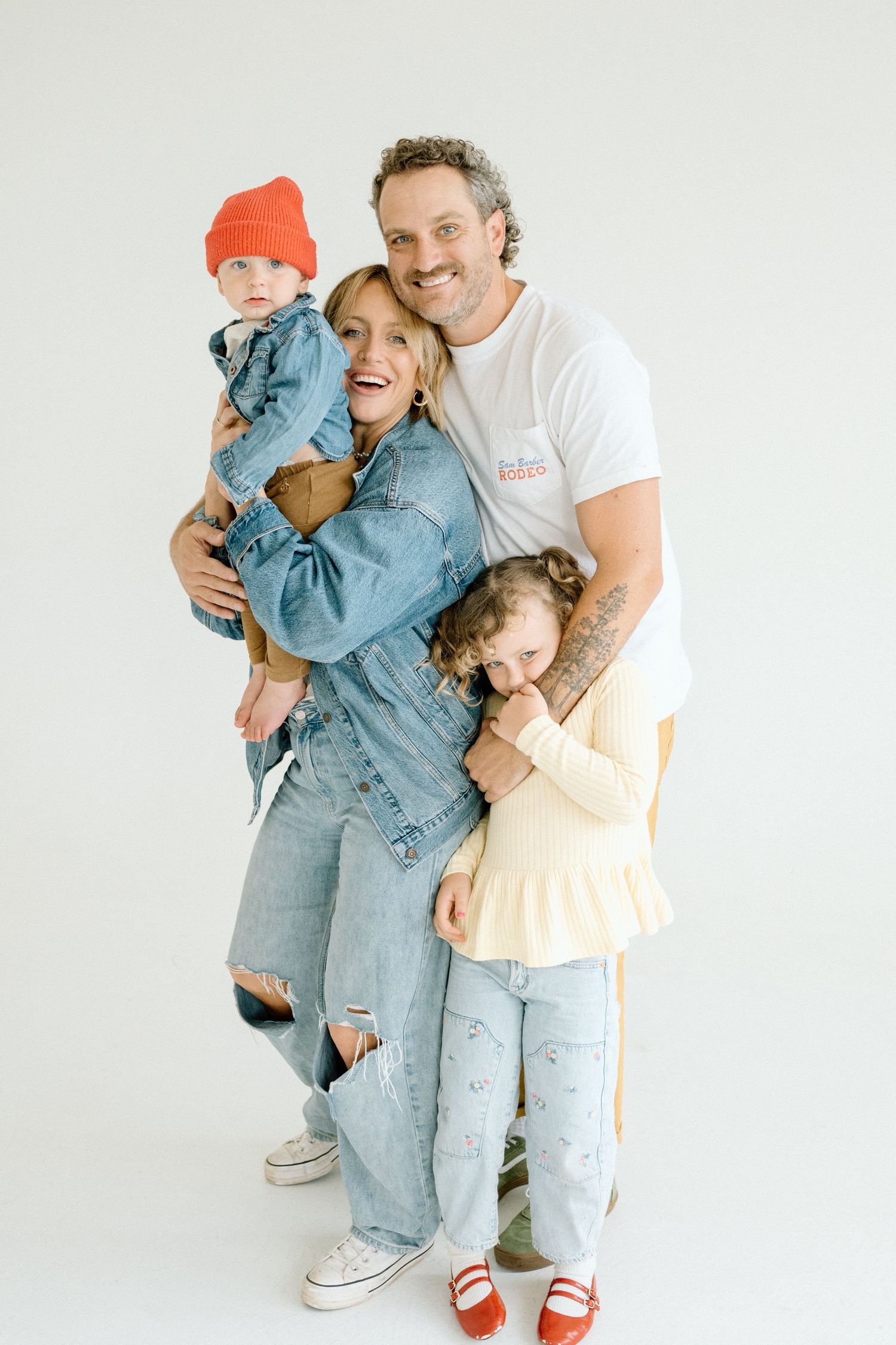 Happy family of four in denim outfits posing together, parents holding young children against white background.