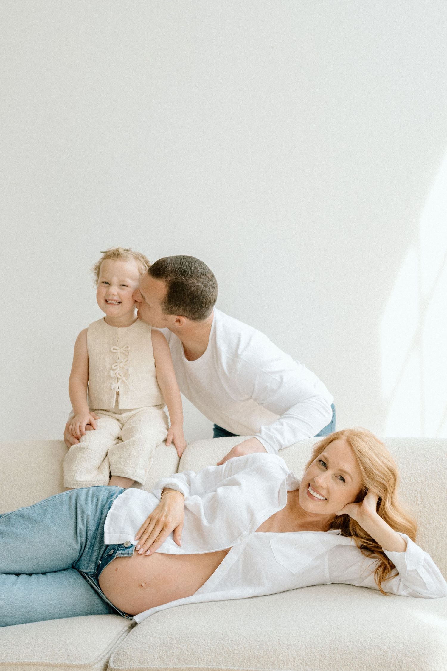 Happy family with pregnant mother, father, and young child posing on white couch.