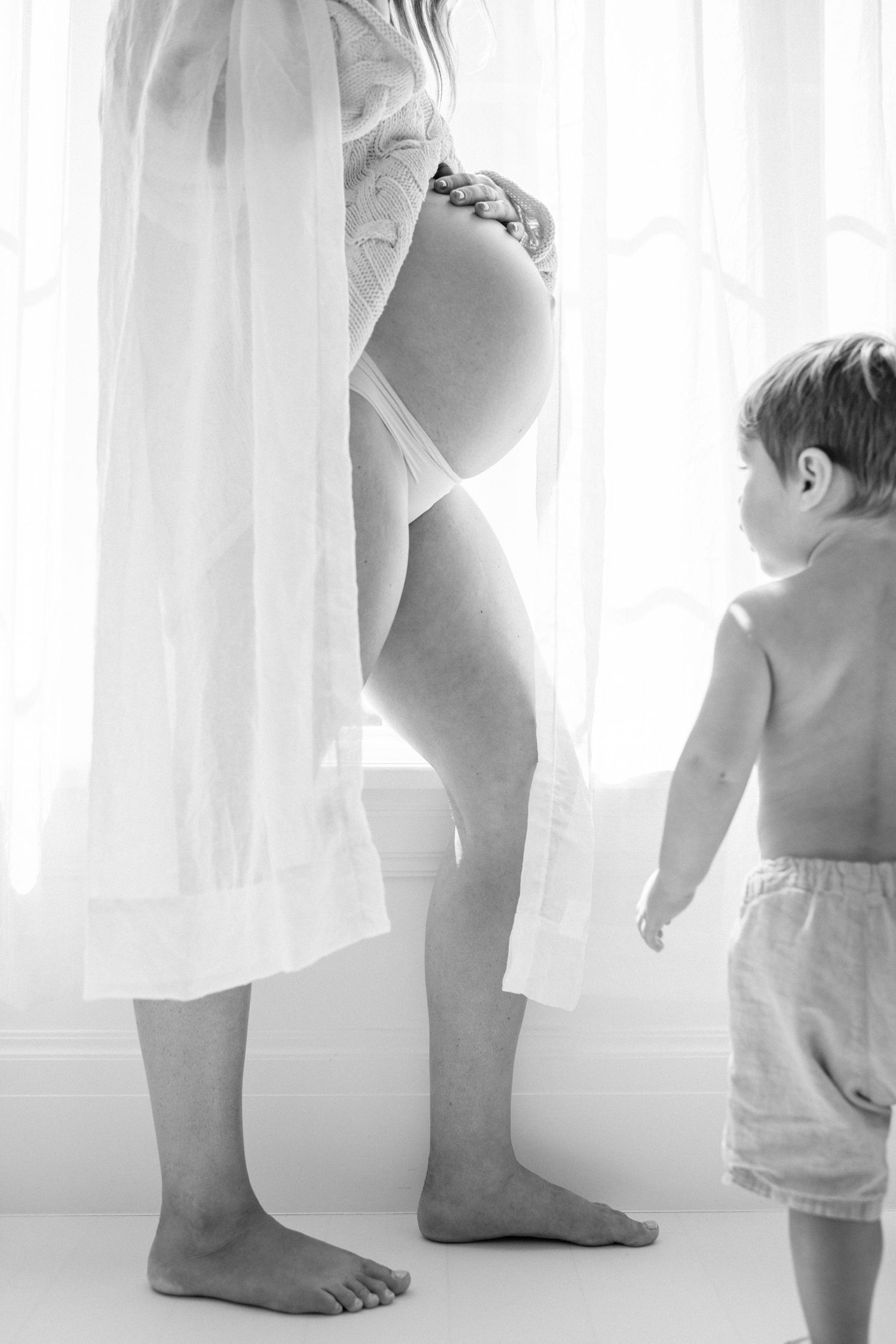 Pregnant woman in white robe stands barefoot near window as toddler boy looks up at her baby bump.