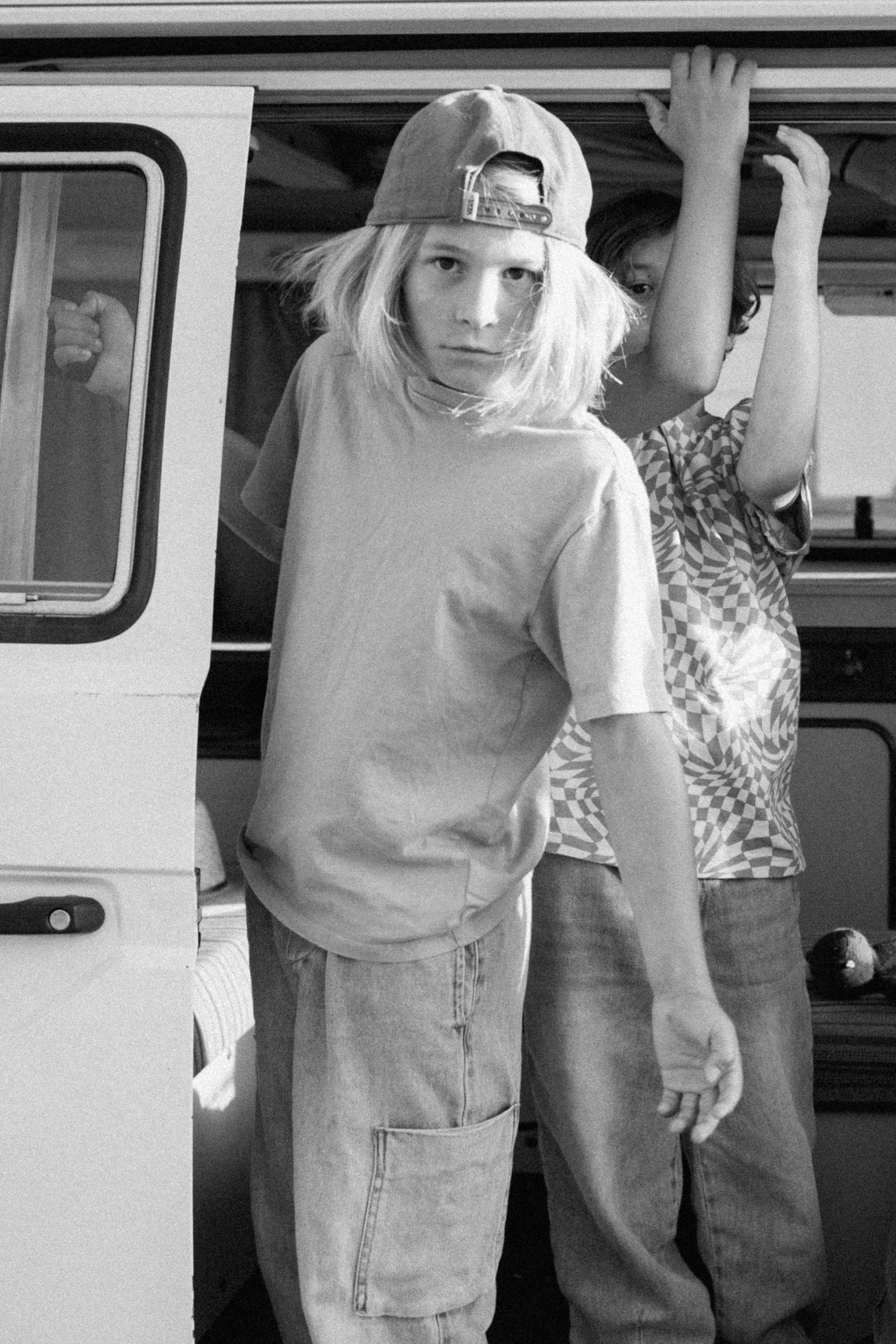 Blonde teenage boy in cap and baggy jeans stands by a white van in black and white photo.