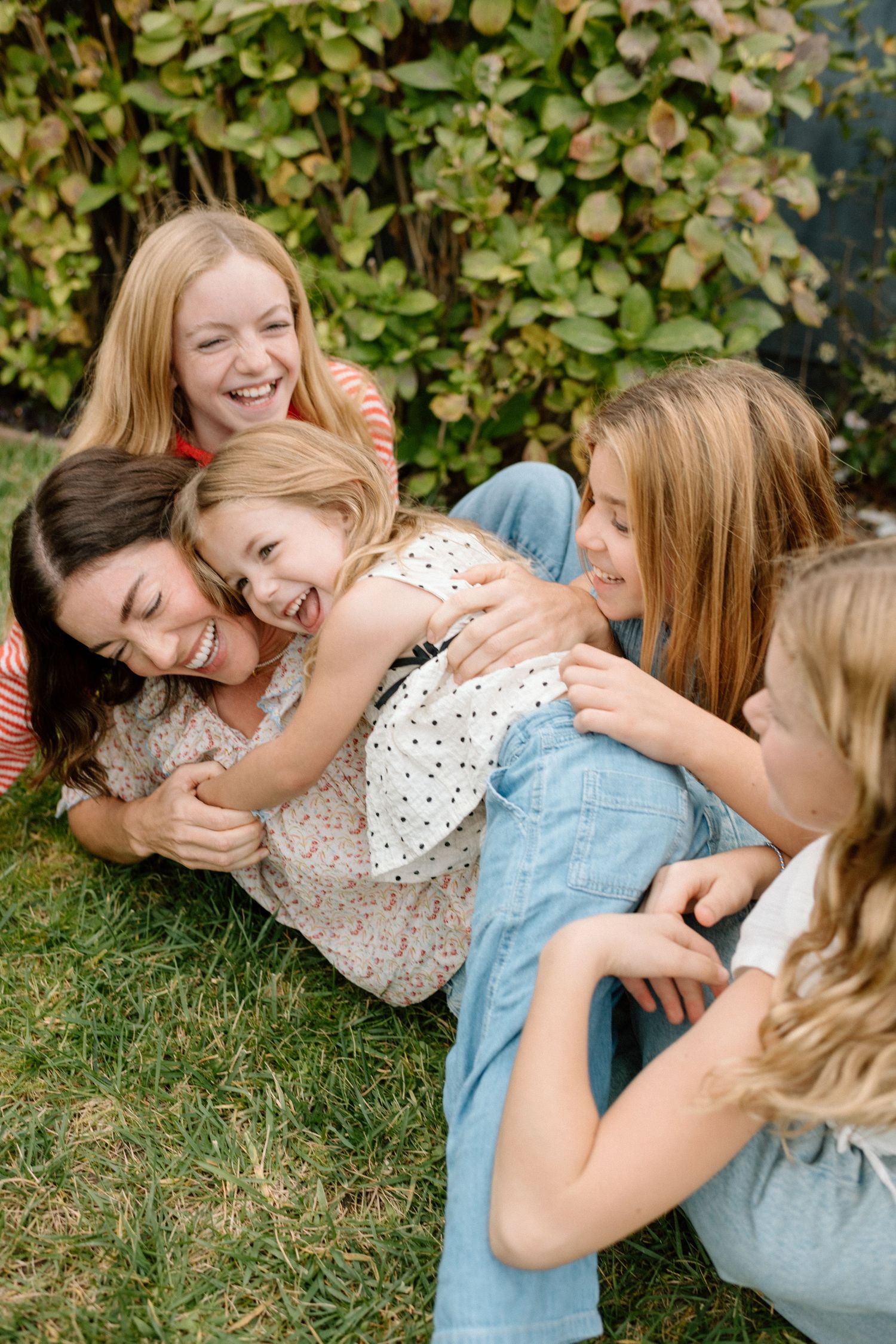 A joyful mother and four daughters laughing and cuddling together on grass in front of lush green bushes.