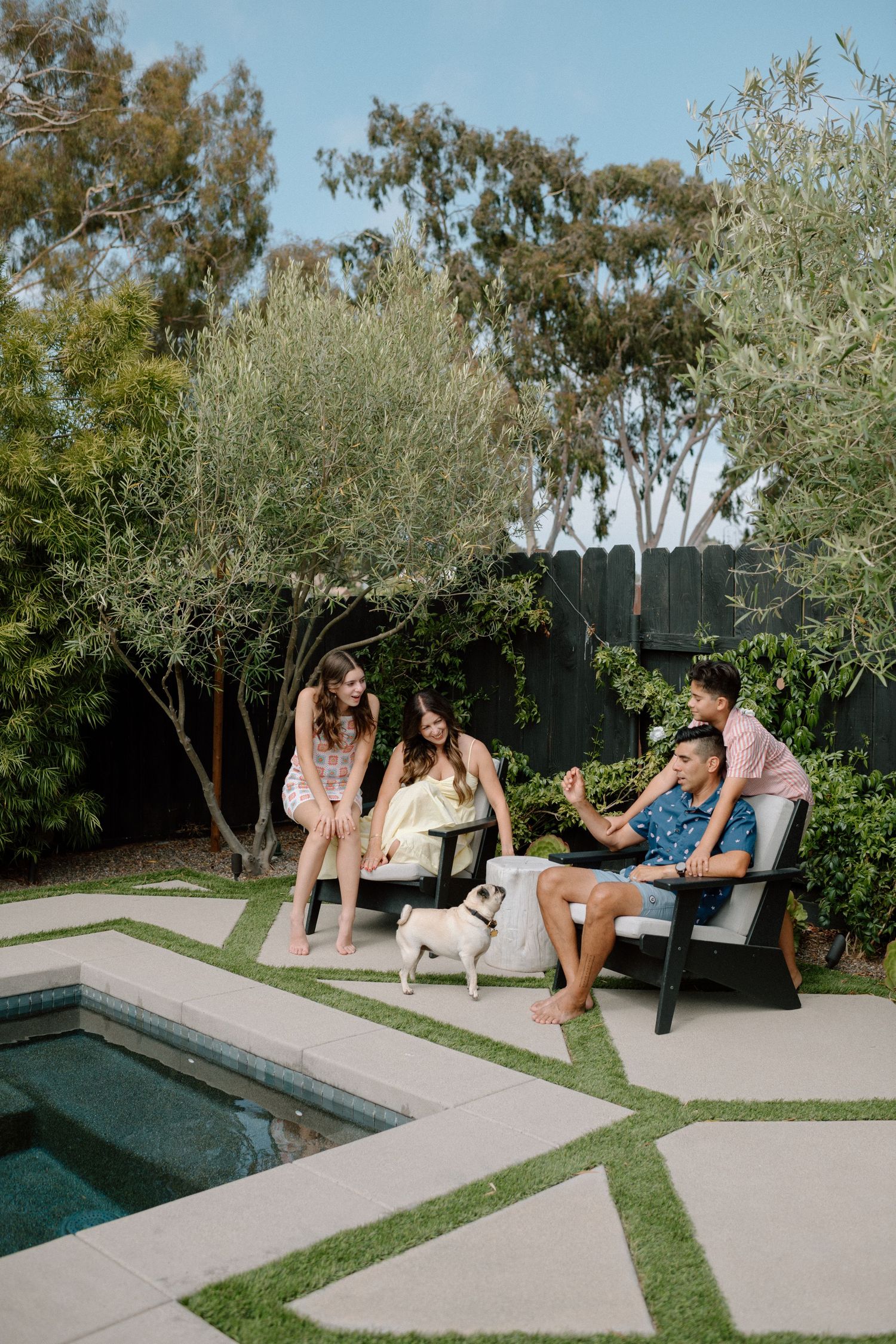 Family of four relaxing outdoors by a pool with a small pug dog on a sunny day.