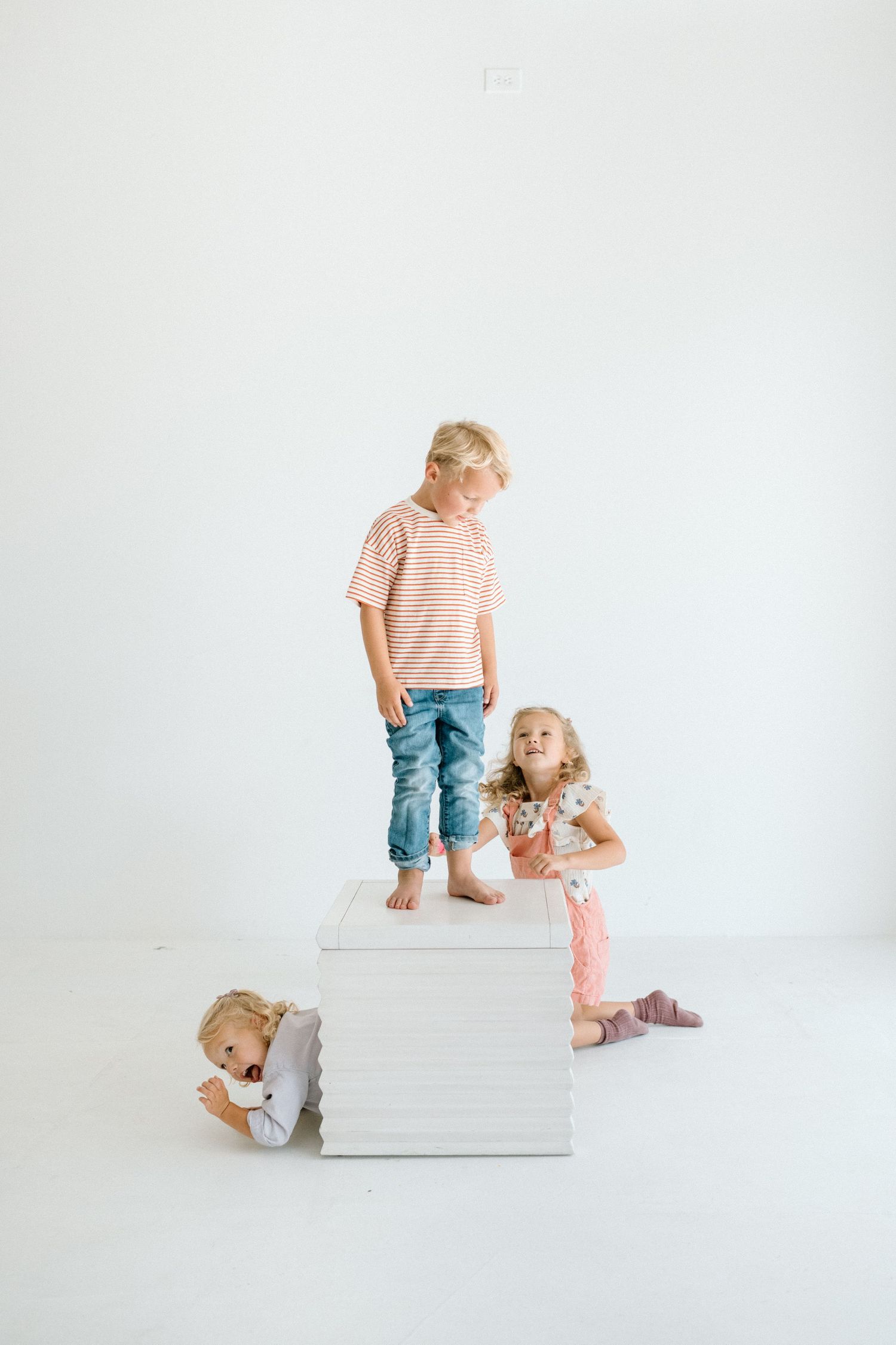 Boy stands on white cube while two young girls play around it in a bright white studio.