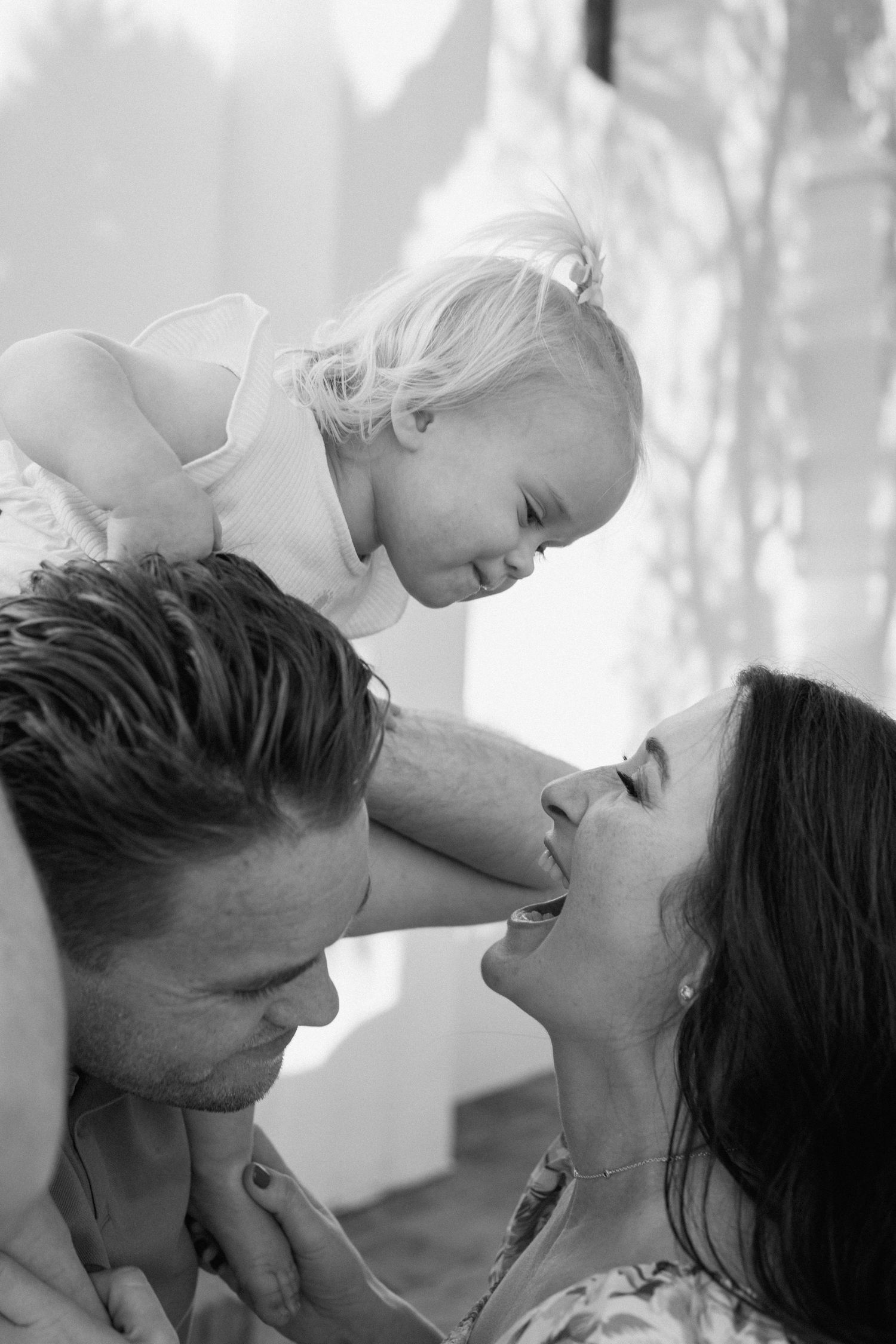 Black and white photo of parents lovingly lifting their laughing baby girl between them indoors.