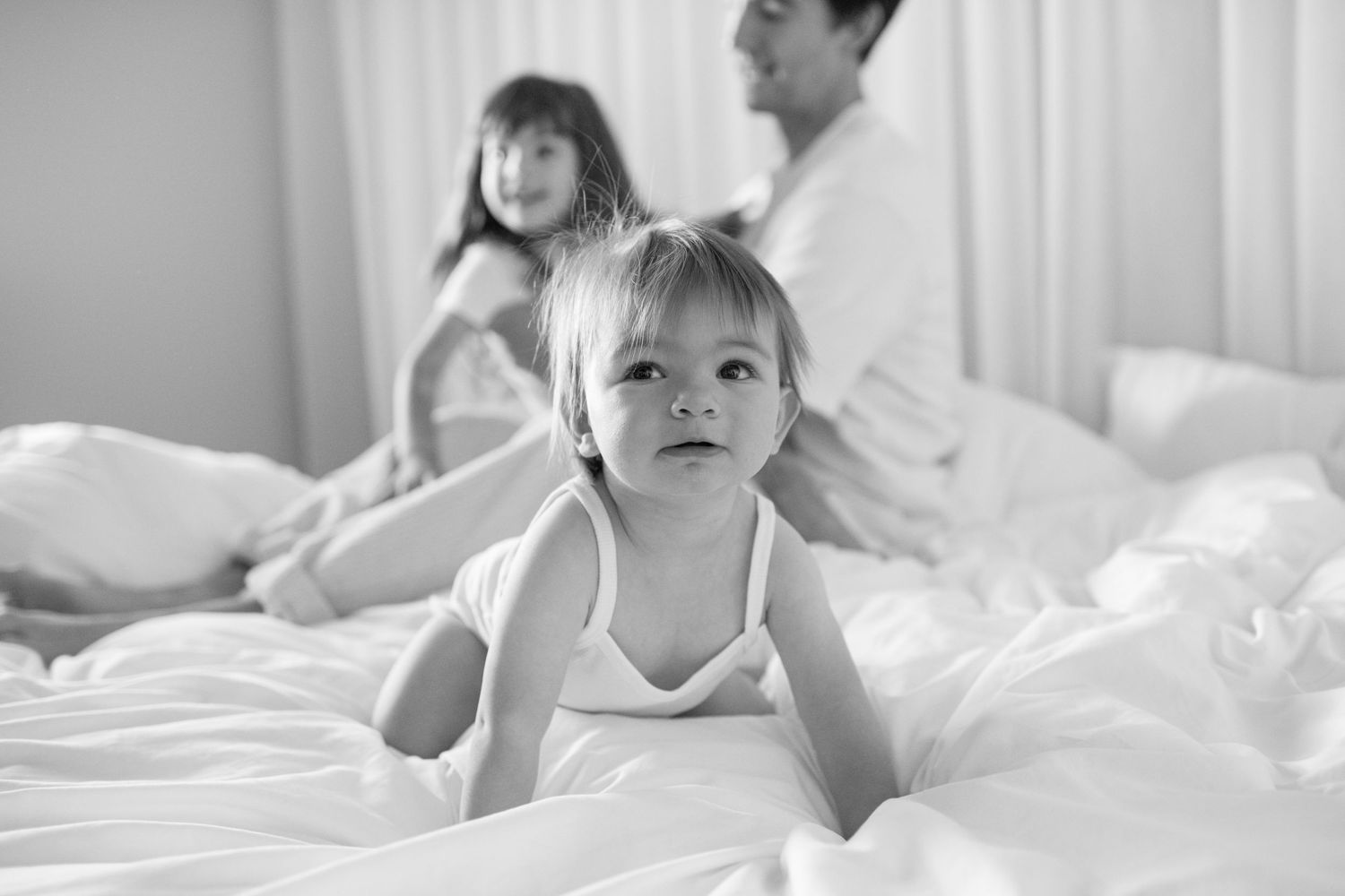 Black and white photo of a toddler on a bed with parents sitting behind, capturing a candid family moment.