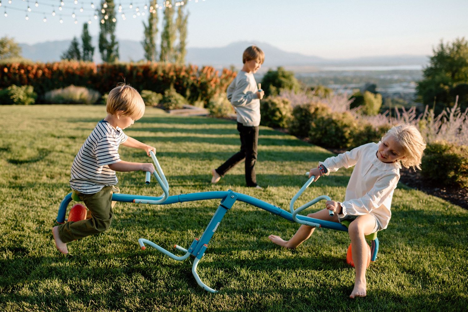 Three children playing on a blue seesaw in a sunny green backyard with scenic hills in the background.