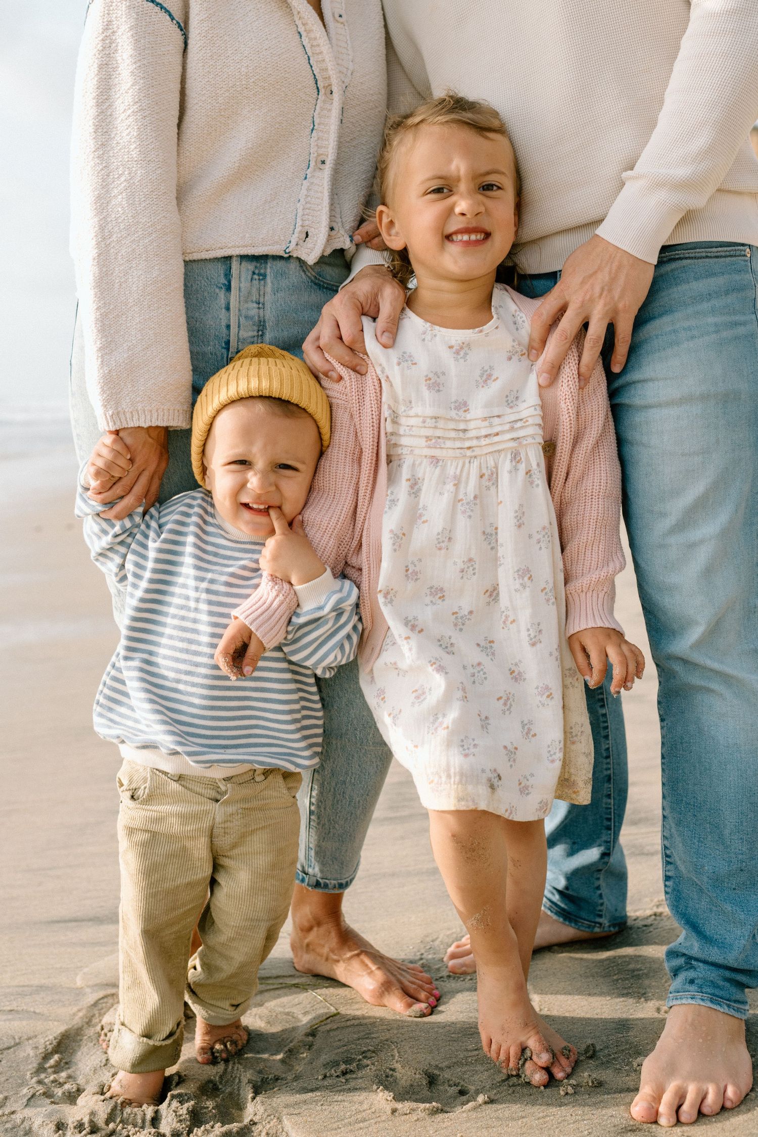 Two young children stand barefoot on sandy beach with parents, girl in white dress and boy in striped shirt wearing yellow hat.
