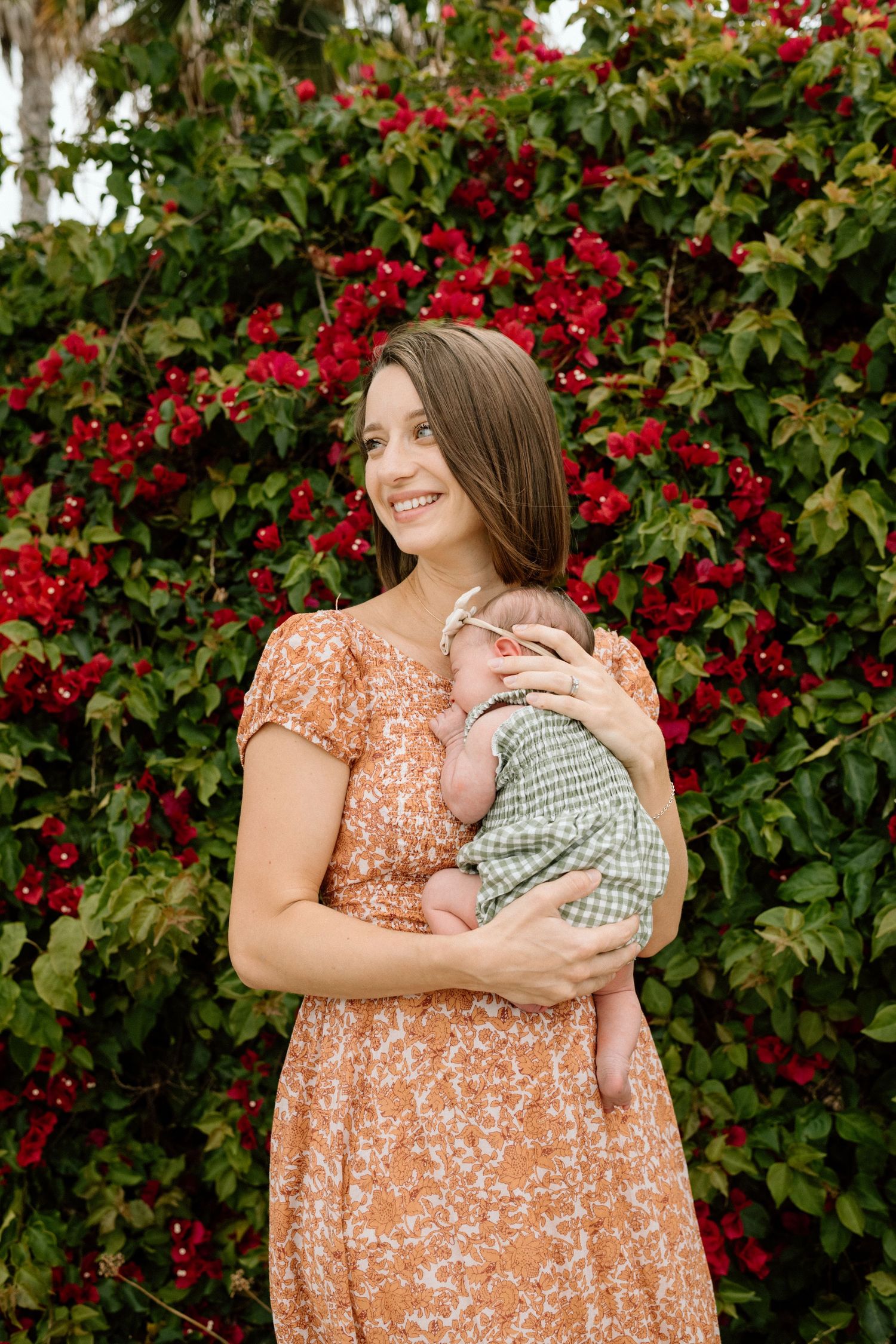 Smiling woman in floral dress holding newborn baby in green outfit against red bougainvillea background.