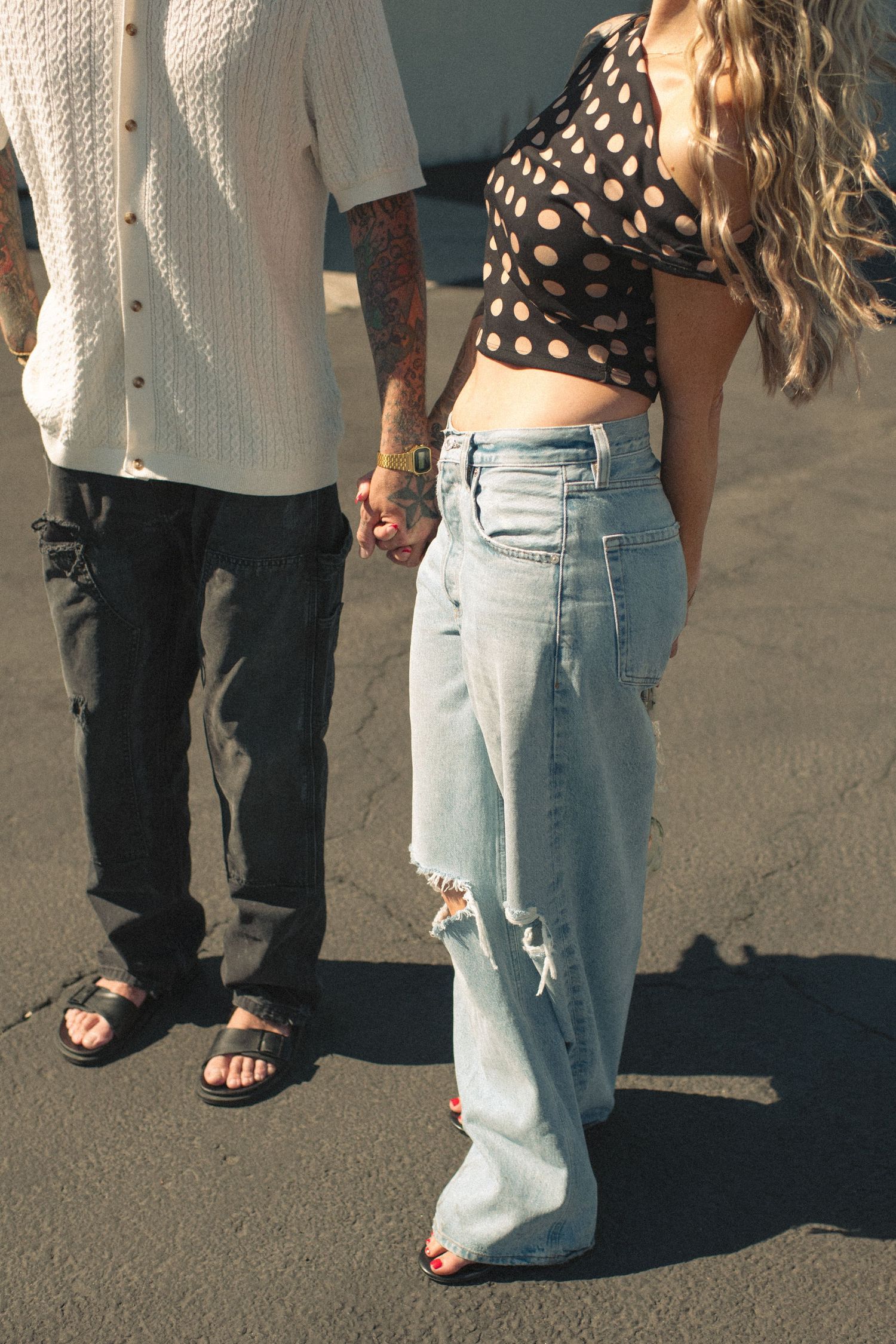 A couple holds hands while walking, the woman wearing ripped wide-leg jeans and a polka dot crop top.