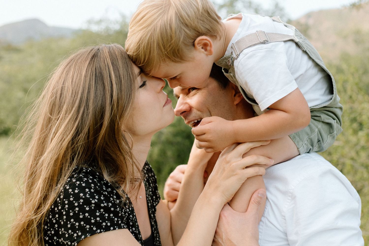 Parents smiling face to face with young child on fathers shoulders in a natural outdoor setting.