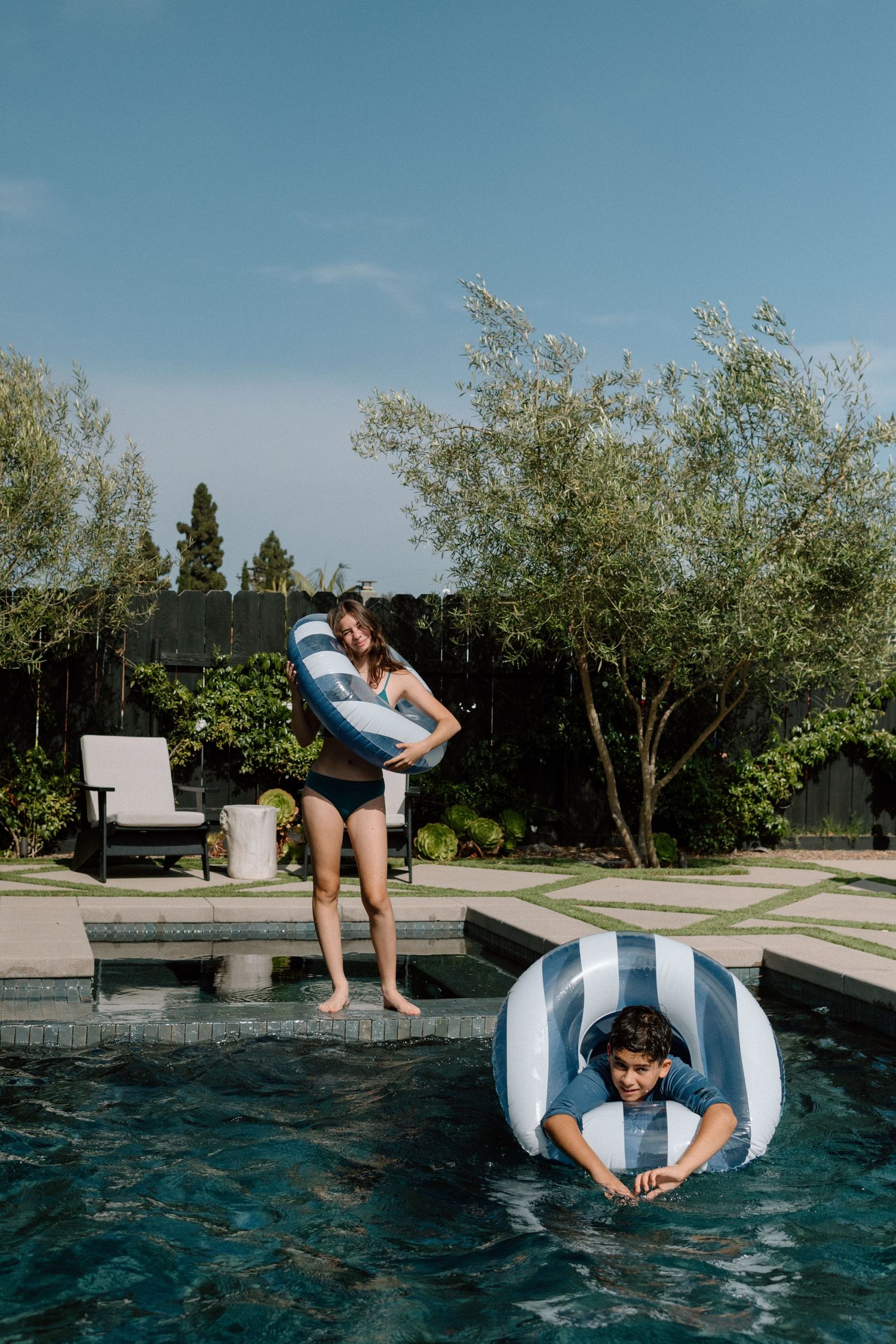 Woman standing poolside holding inflatable ring while boy floats in striped pool float.