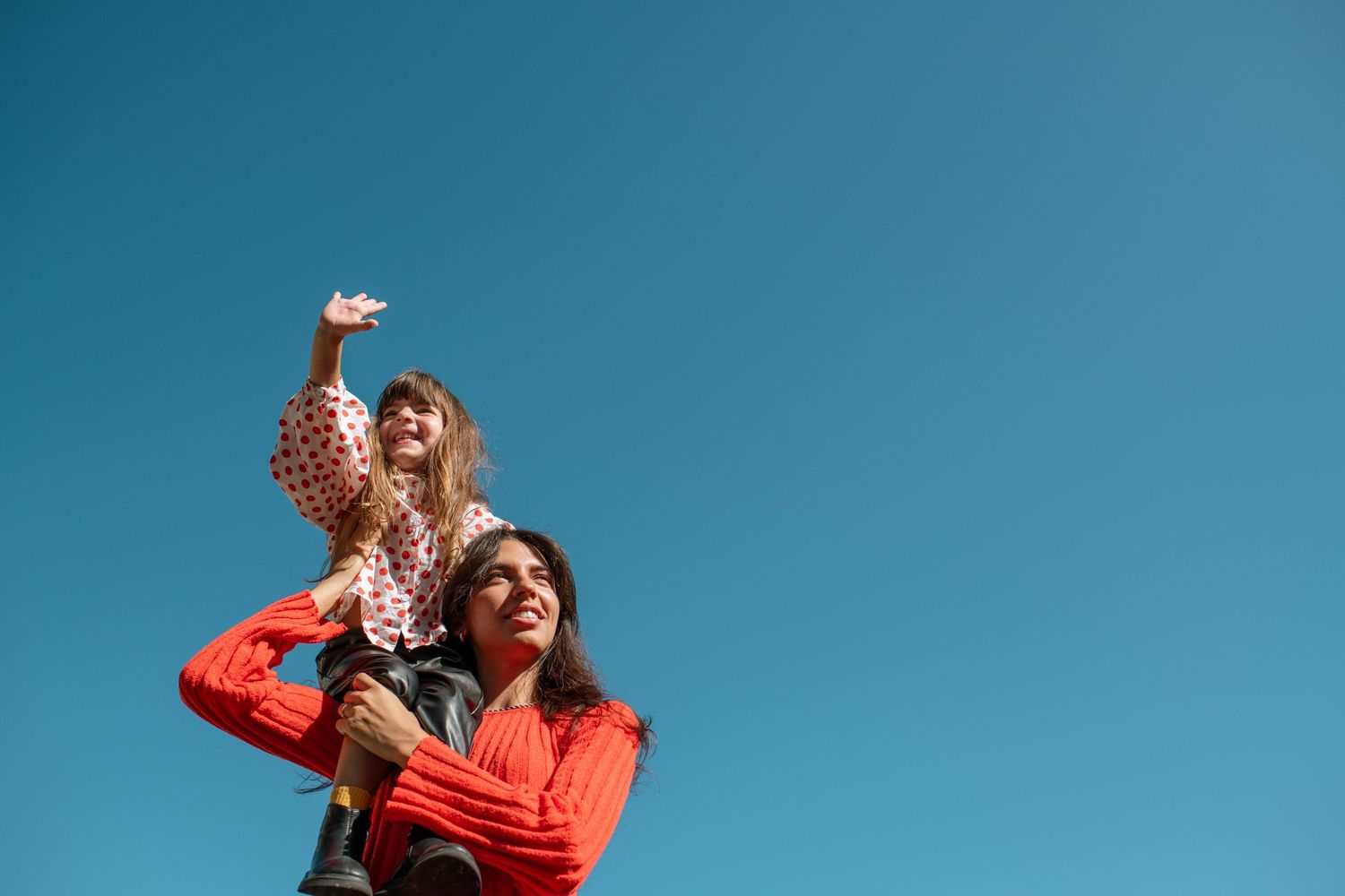A joyful child sits on a woman's shoulders against a clear blue sky, both wearing red, as the child reaches up happily.