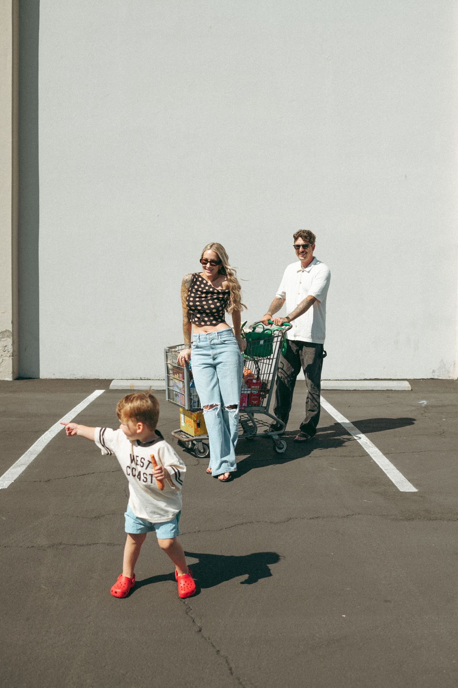 Young family pushing full grocery cart in parking lot, stylish mom in sunglasses, dad smiling, toddler walking ahead.