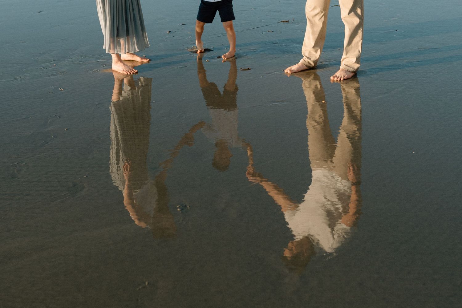 Three people stand barefoot on a reflective wet beach, their reflections mirrored in the water.