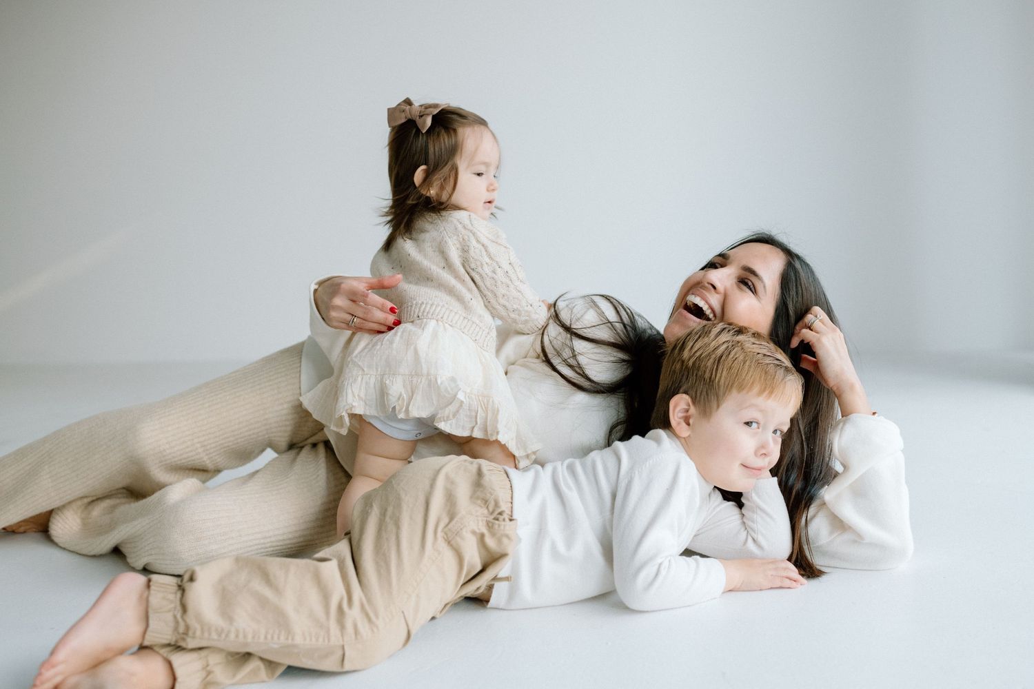 A joyful mother sits on a white floor laughing as her two young children playfully climb on her.