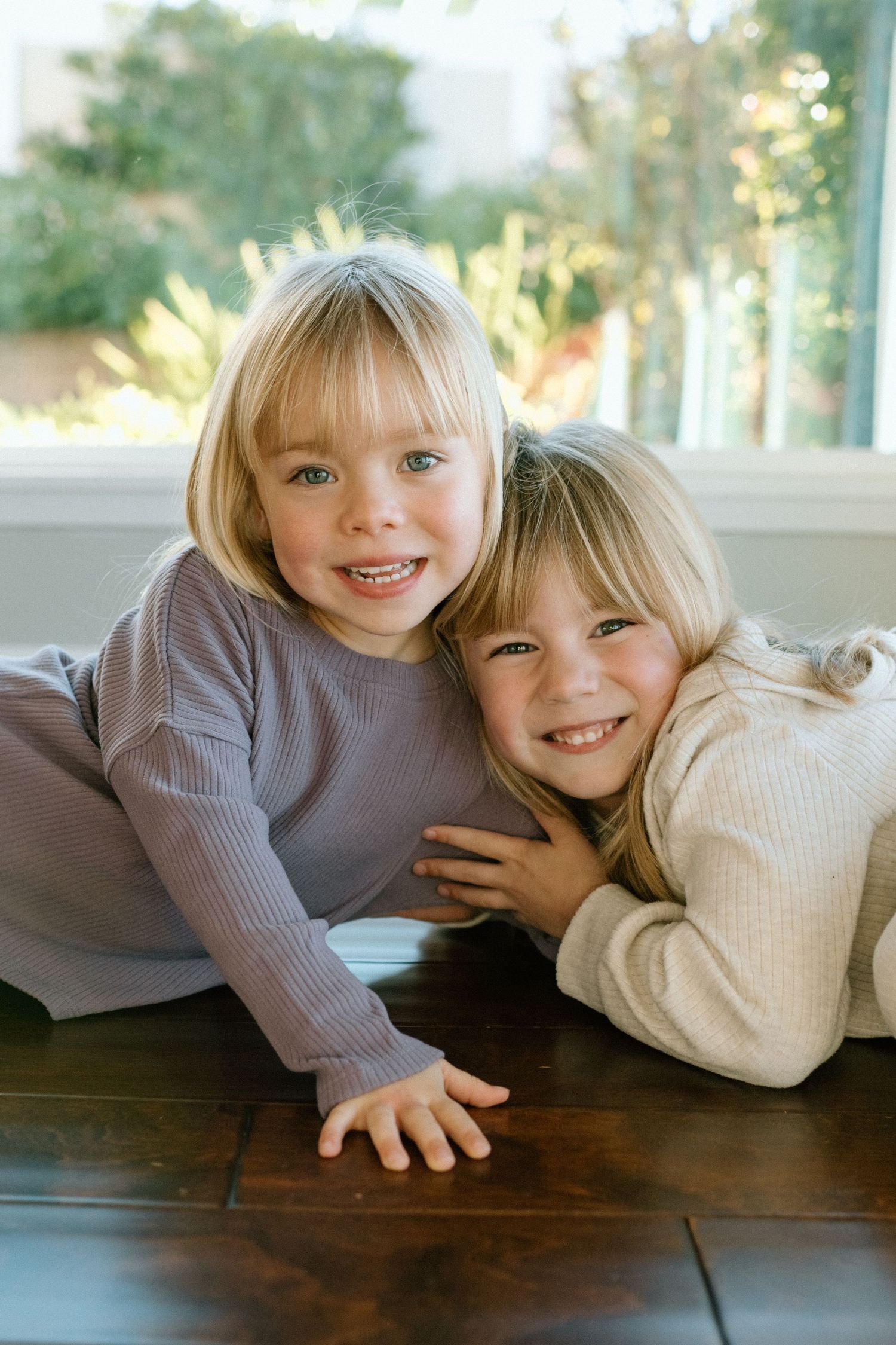 Two smiling blonde girls in neutral ribbed tops lying on hardwood floor near sunlit window.