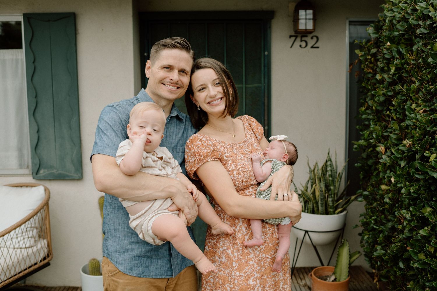 Happy young family of four posing outdoors, parents holding two babies in front of home entrance numbered 752.