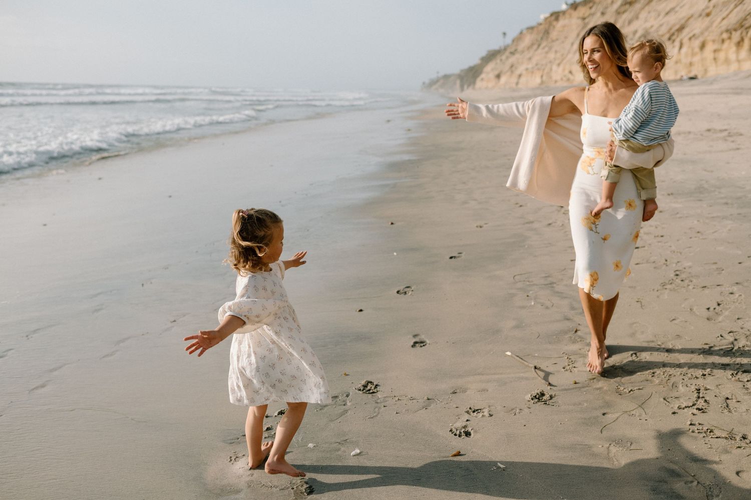 A mother holds a baby while reaching out to her young daughter on a sandy beach near the ocean.