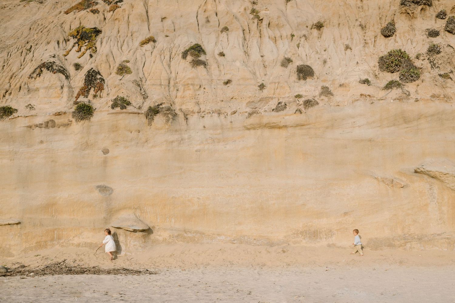 Two people walking on a sandy beach below towering golden sandstone cliffs with sparse vegetation.