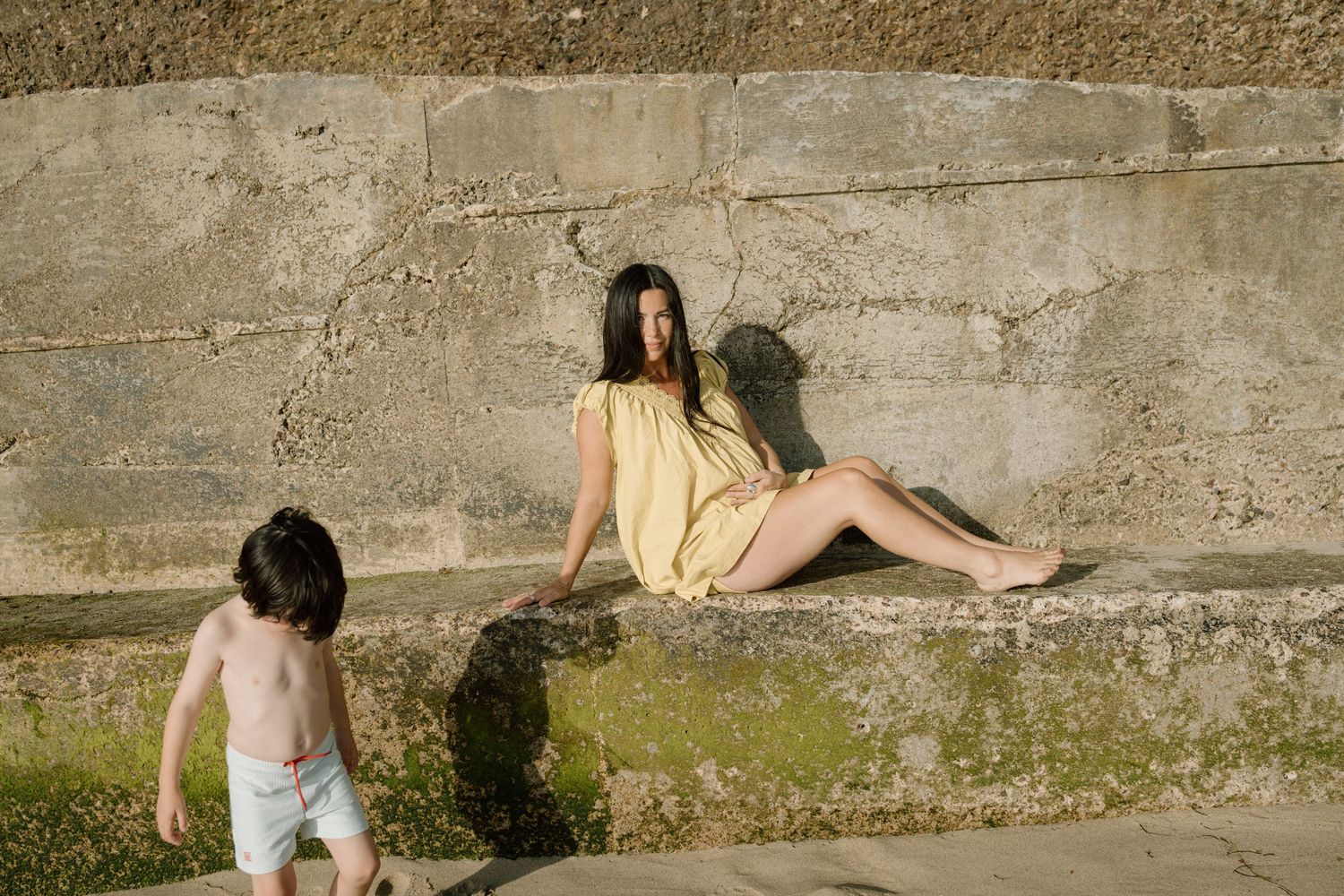 A woman in a yellow dress sits on stone steps while a young child plays nearby outdoors.