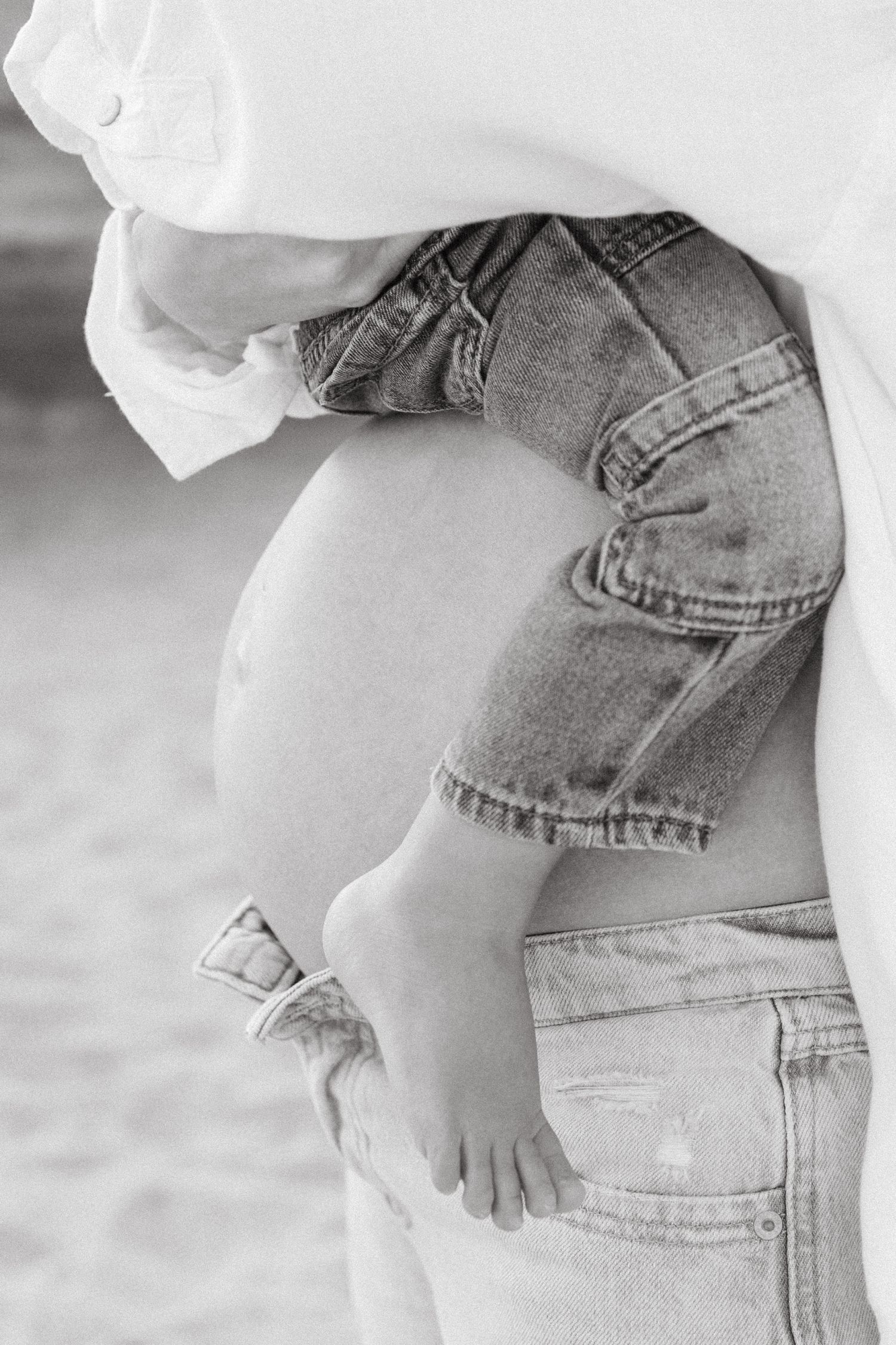 Black and white close-up of a pregnant womans belly next to a baby in denim shorts being held nearby.