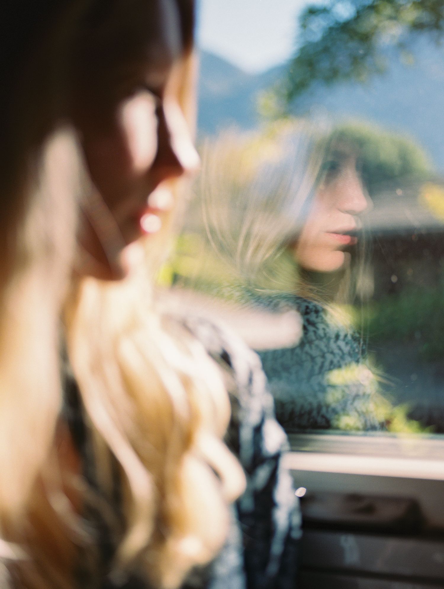 Woman gazes at her reflection in a car window with mountains visible in background.