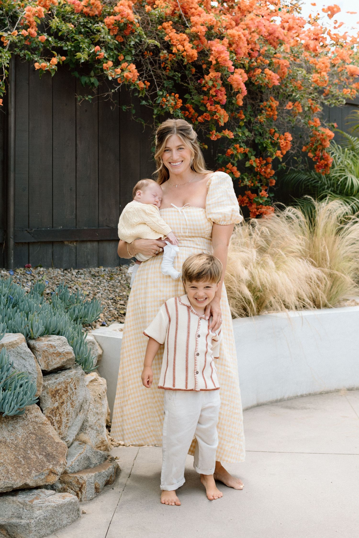 Smiling mother in yellow dress holding newborn baby with toddler son standing outdoors.