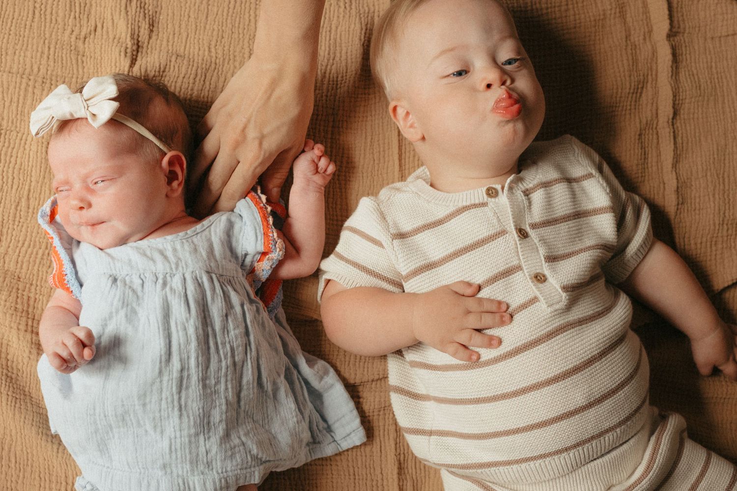 Two babies lying side by side on a tan blanket, one newborn in blue dress with bow, one older infant in striped outfit.
