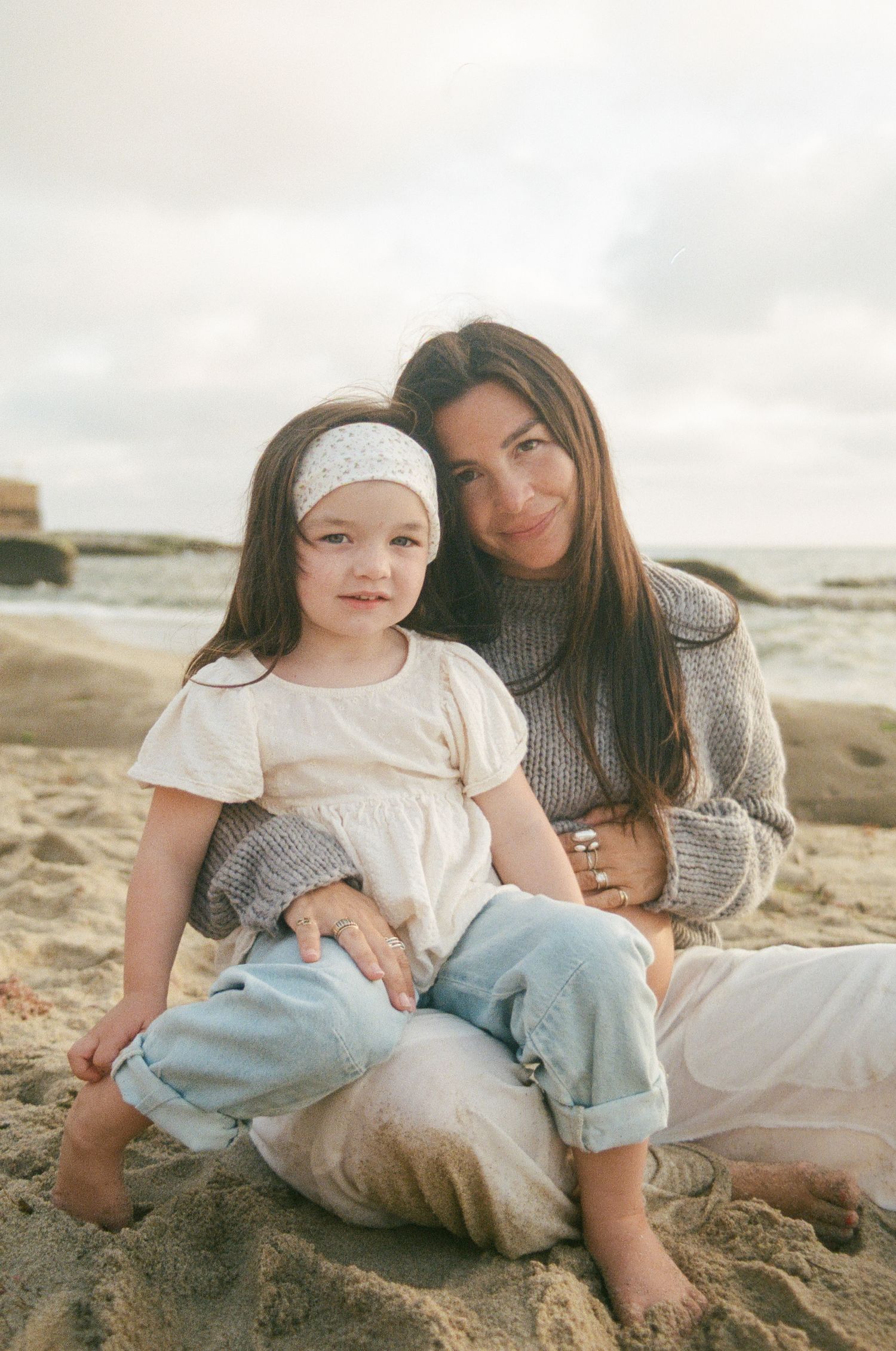 A woman and young girl sit together on sandy beach, the child wearing a white dress and headband.