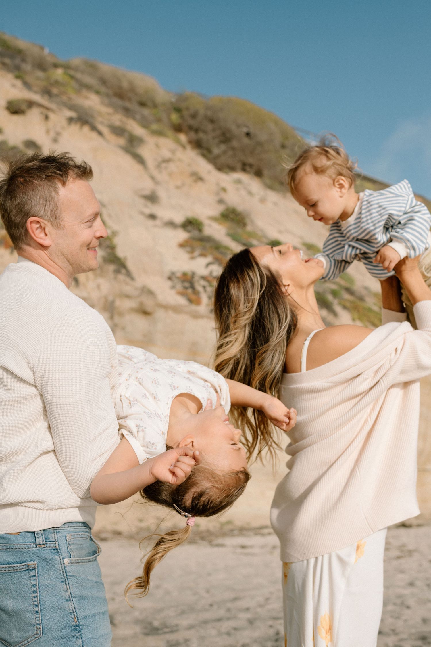 Happy family at beach, parents playfully lifting two young children against sandy cliffs and blue sky.