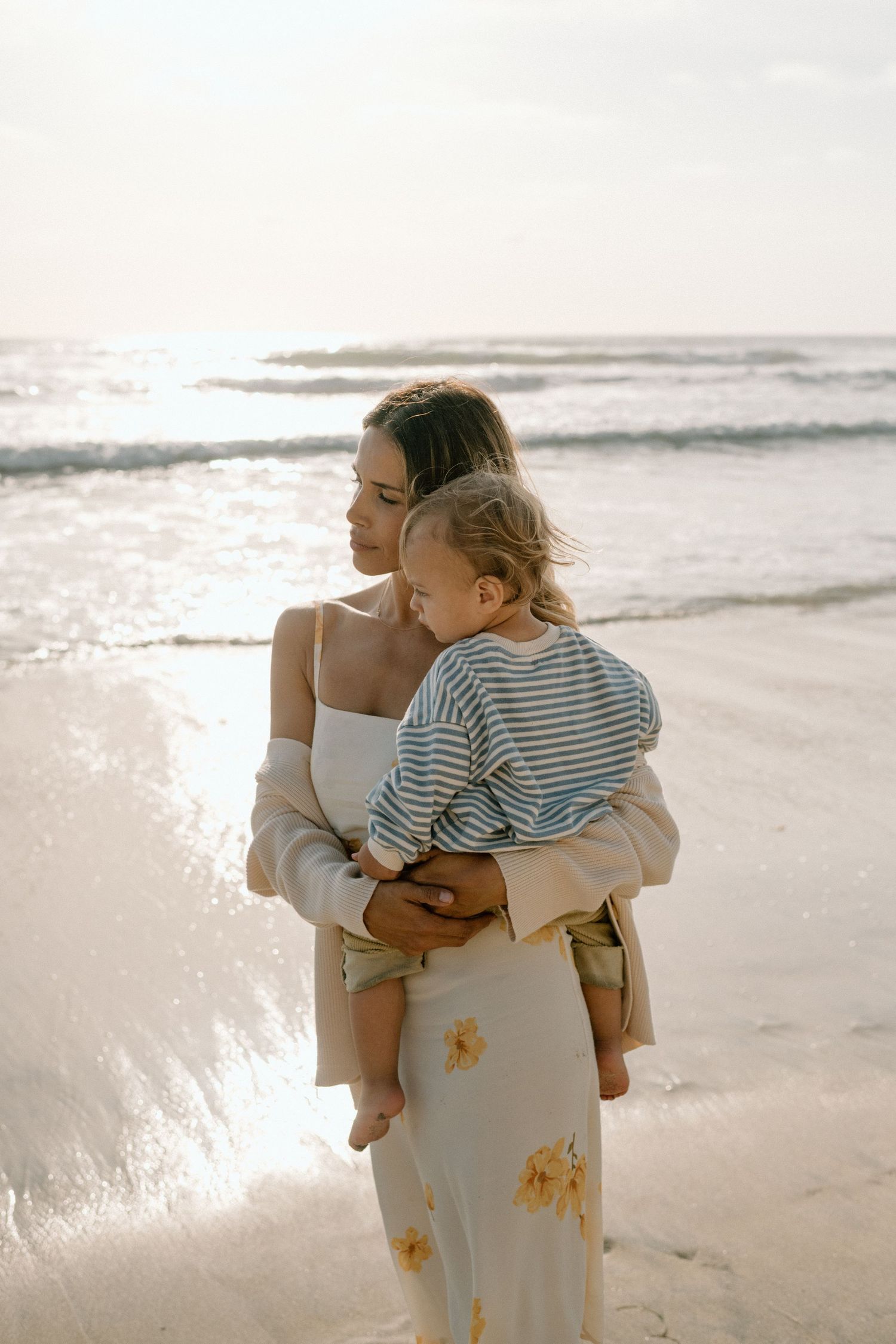 A young mother holds her toddler on a sunlit beach, both gazing toward the ocean waves.