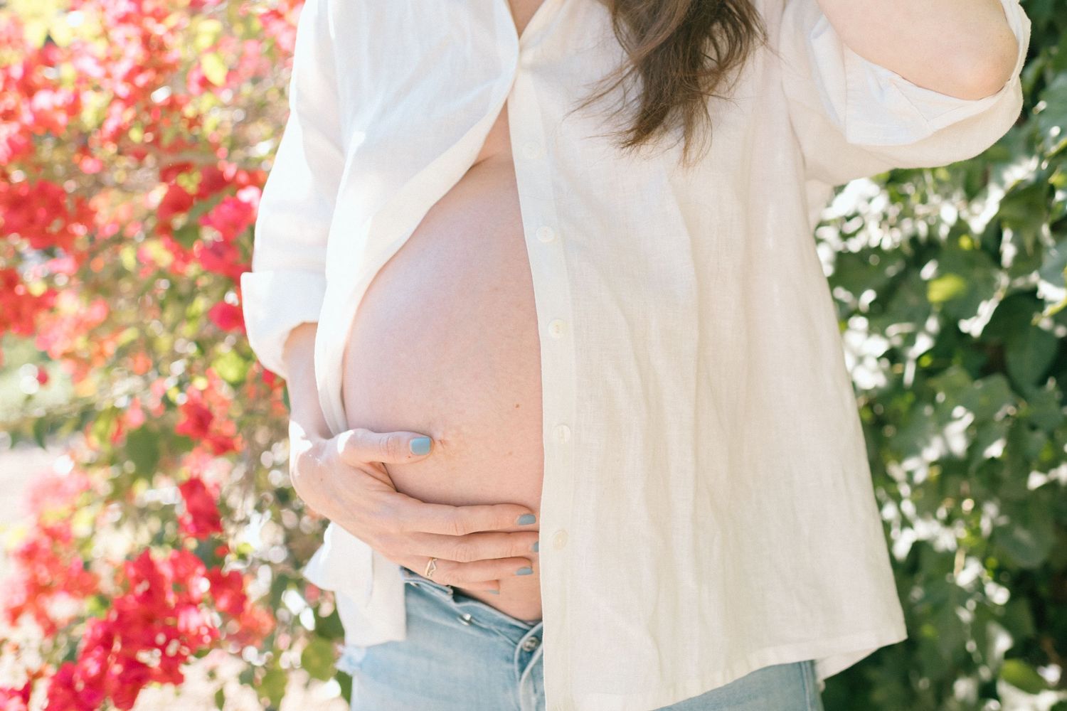 Pregnant woman in white shirt and jeans gently holding her bare baby bump outdoors.
