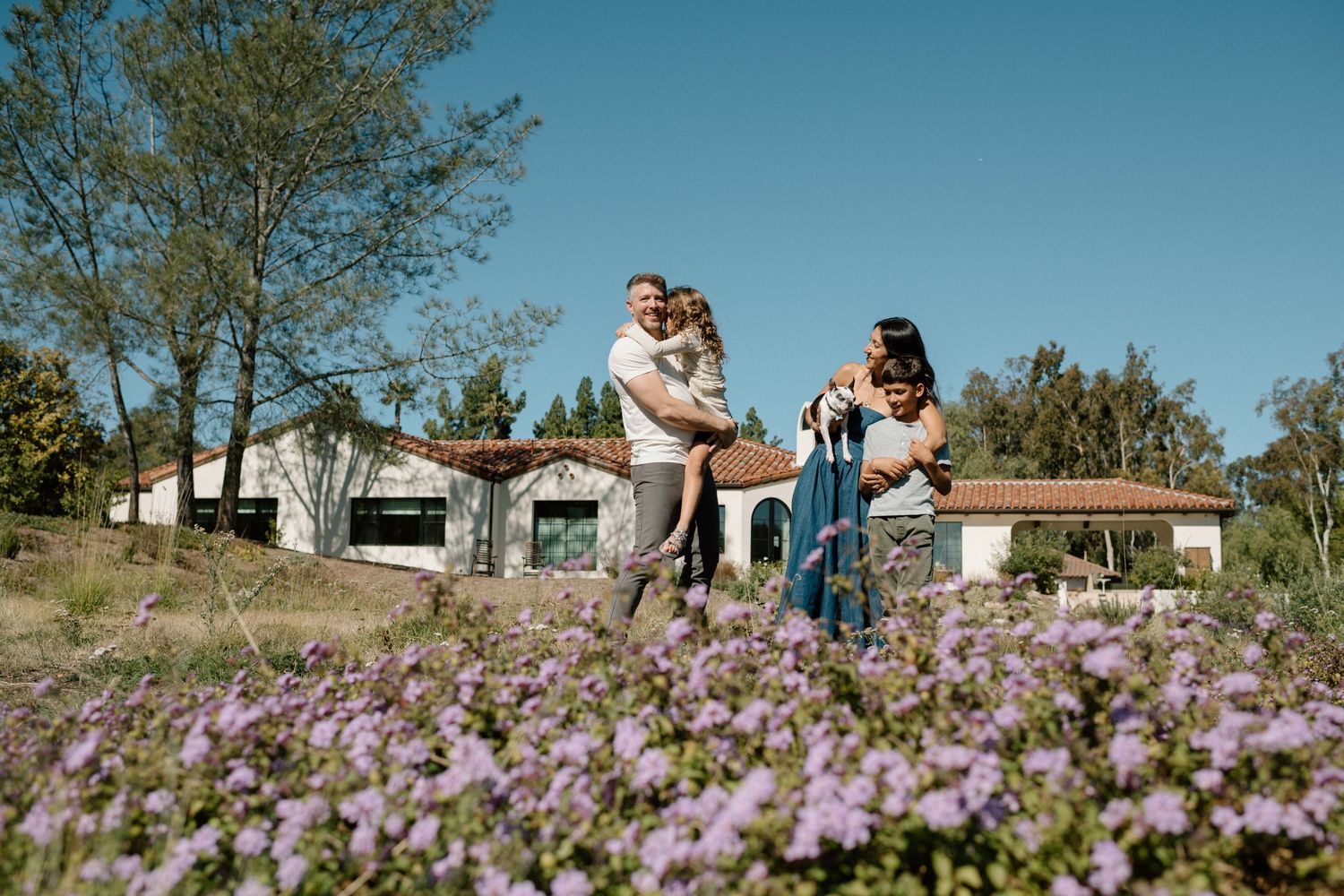 Happy family playing outdoors near a rural home surrounded by purple wildflowers under a clear blue sky.