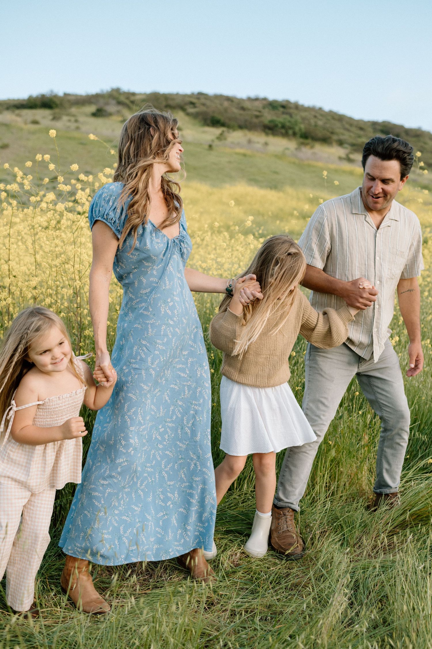 A joyful family walks through a sunny wildflower meadow, with mom in a blue floral dress holding hands with two blonde girls.