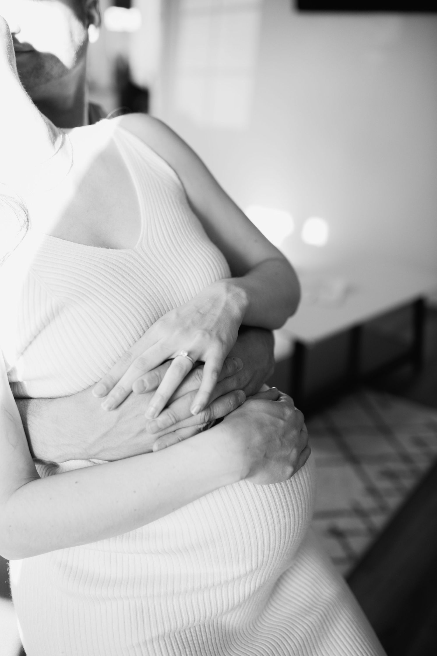 Black and white close-up of partner embracing pregnant woman in white ribbed dress.