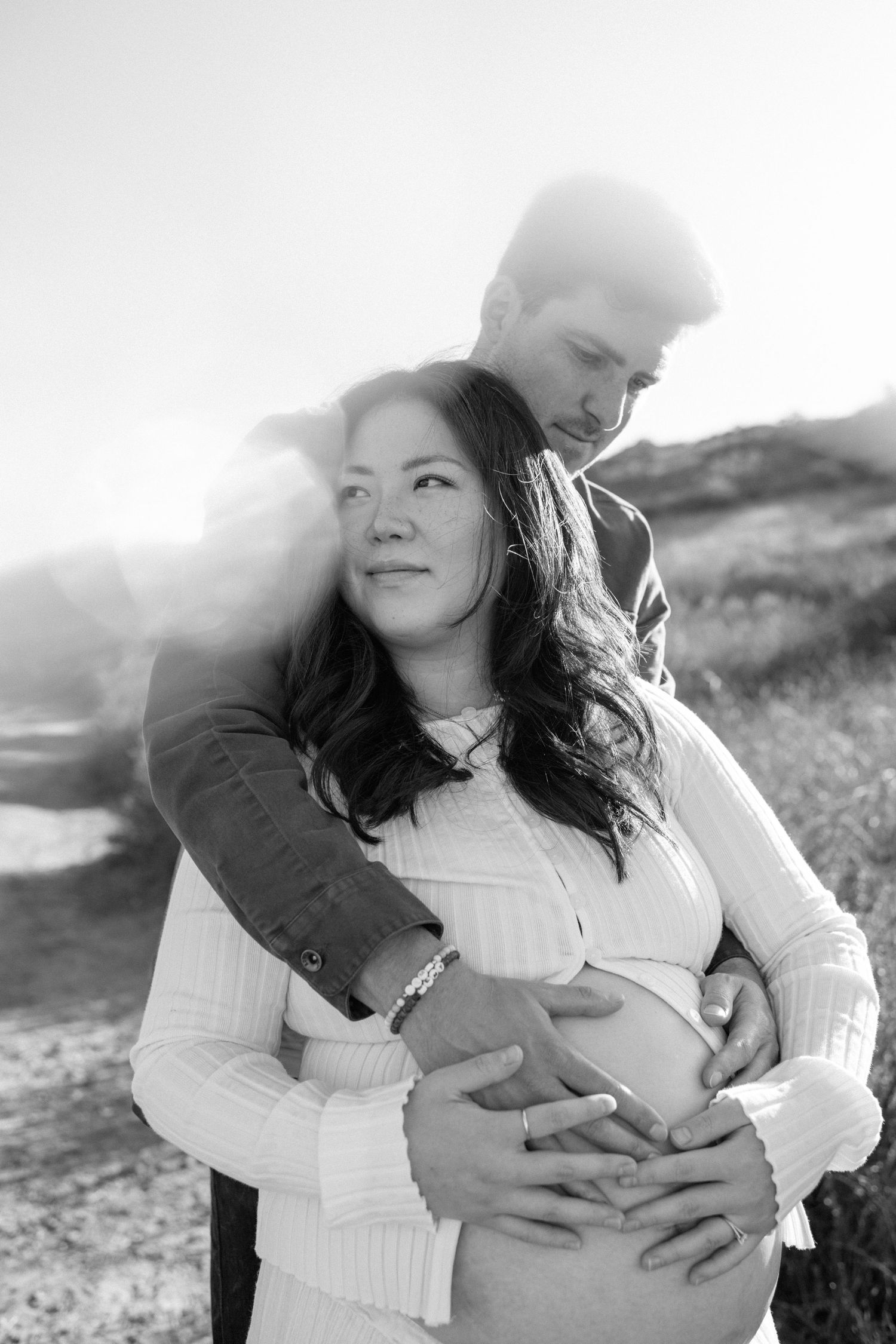 Black and white maternity photo of couple embracing outdoors, man holding pregnant womans belly.