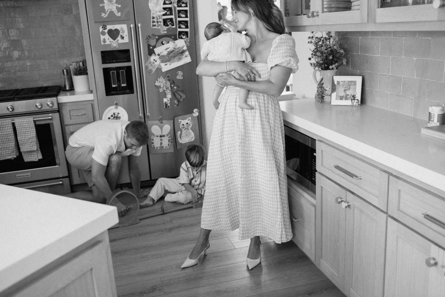 Black and white photo of a mother holding a newborn in a kitchen while two young children play on the floor.