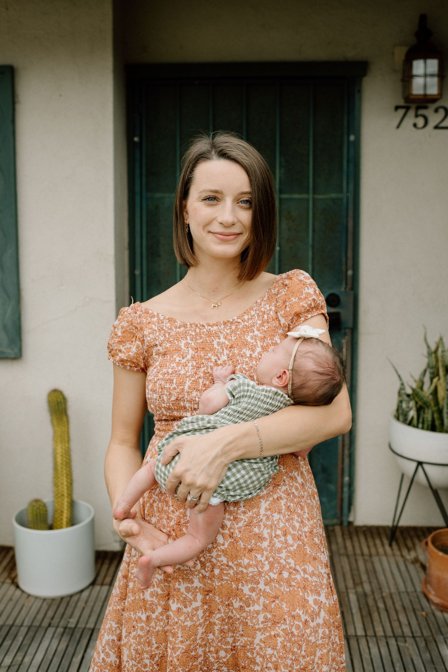 Young mother in floral orange dress holding sleeping newborn baby outside home entrance.